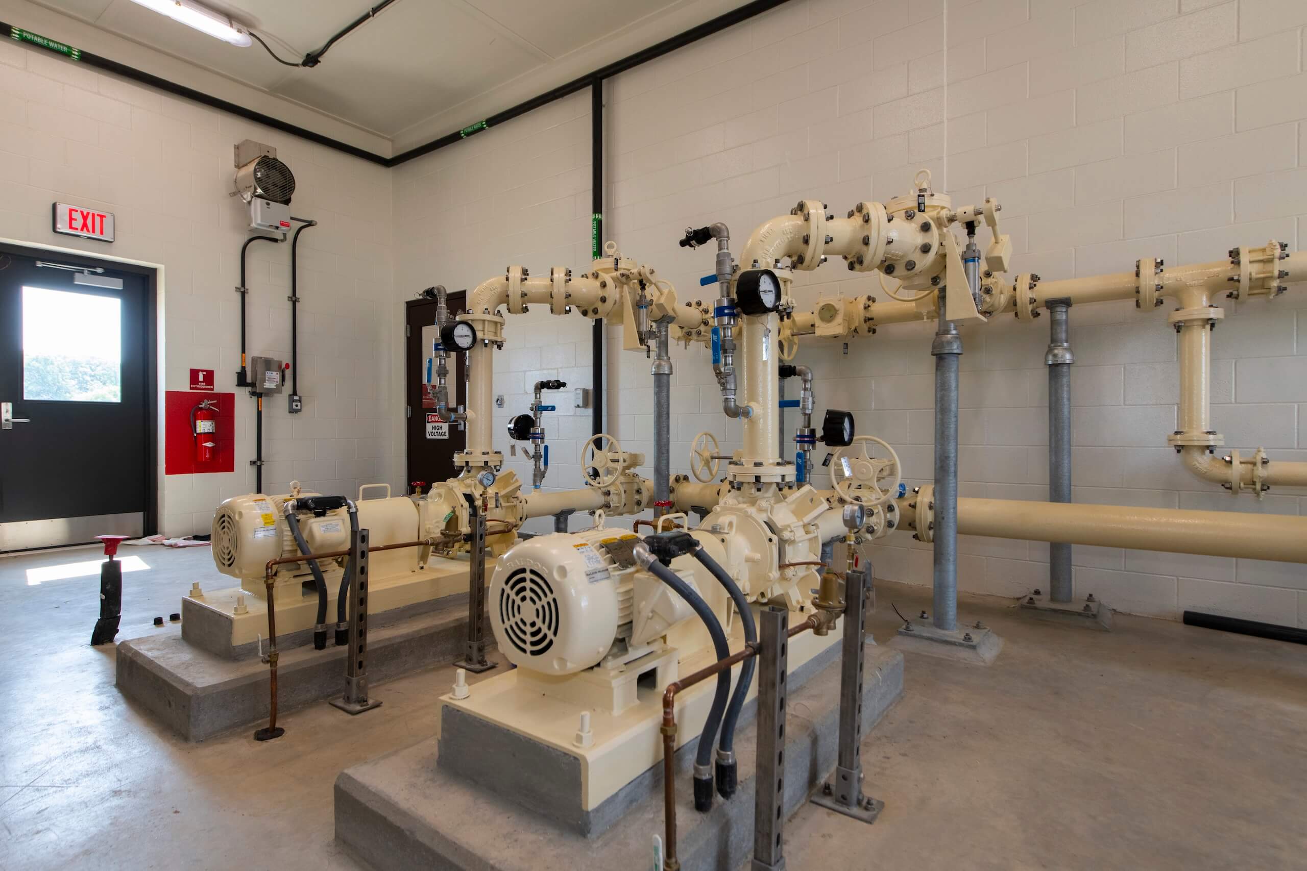 Two large industrial pumps and yellow pipes are installed in a clean, well-lit room with white walls, a concrete floor, and a black door. Pressure gauges and safety devices are attached to the piping system.