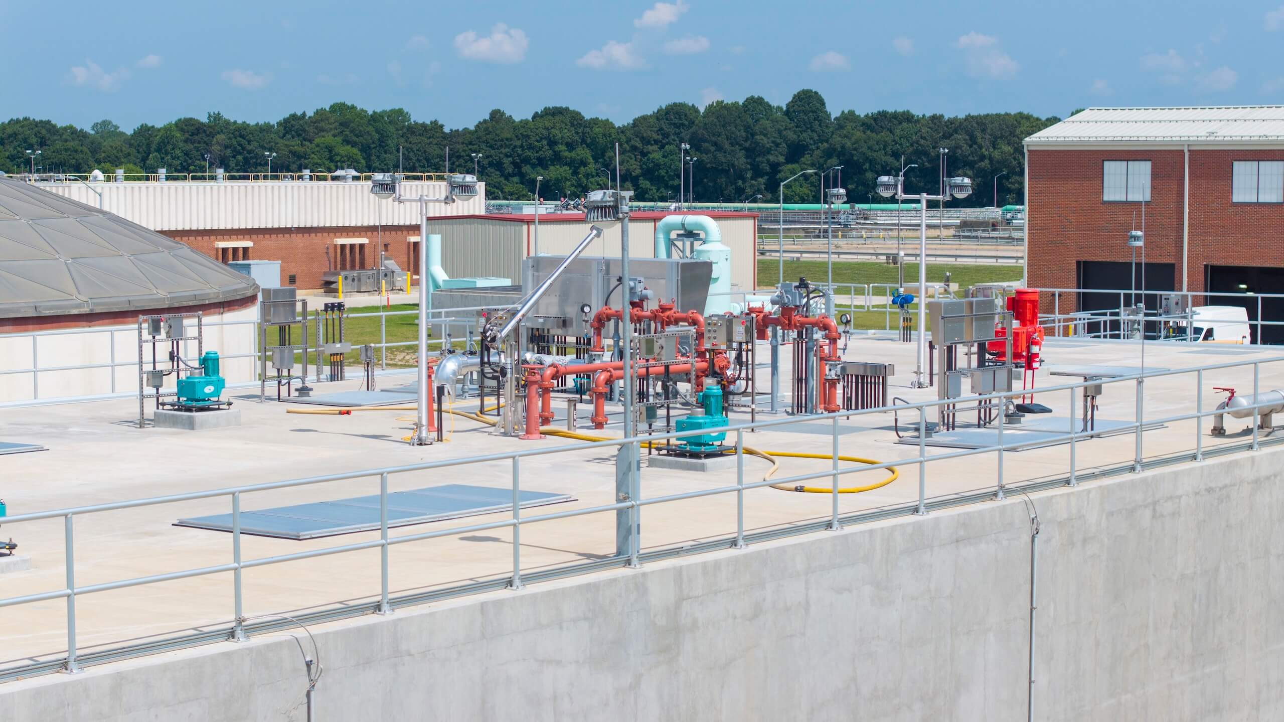 A water treatment facility with various pipes, pumps, tanks, and equipment on a concrete platform; brick buildings and trees are visible in the background under a clear sky.