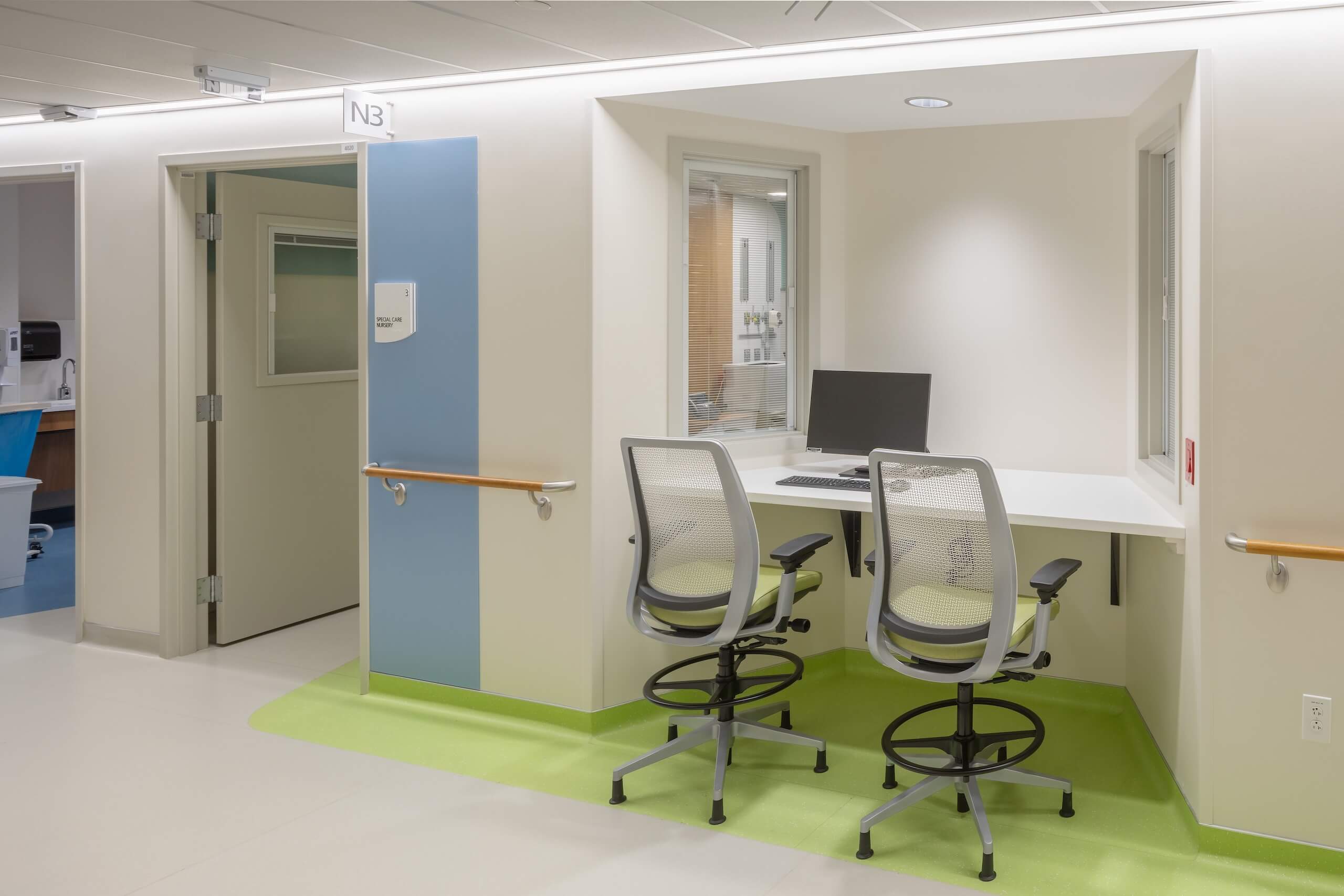 A hospital workstation with two mesh office chairs, a computer on a white desk, and windows, set on a green floor mat. Nearby, a doorway leads to another room. The area is clean and well-lit.