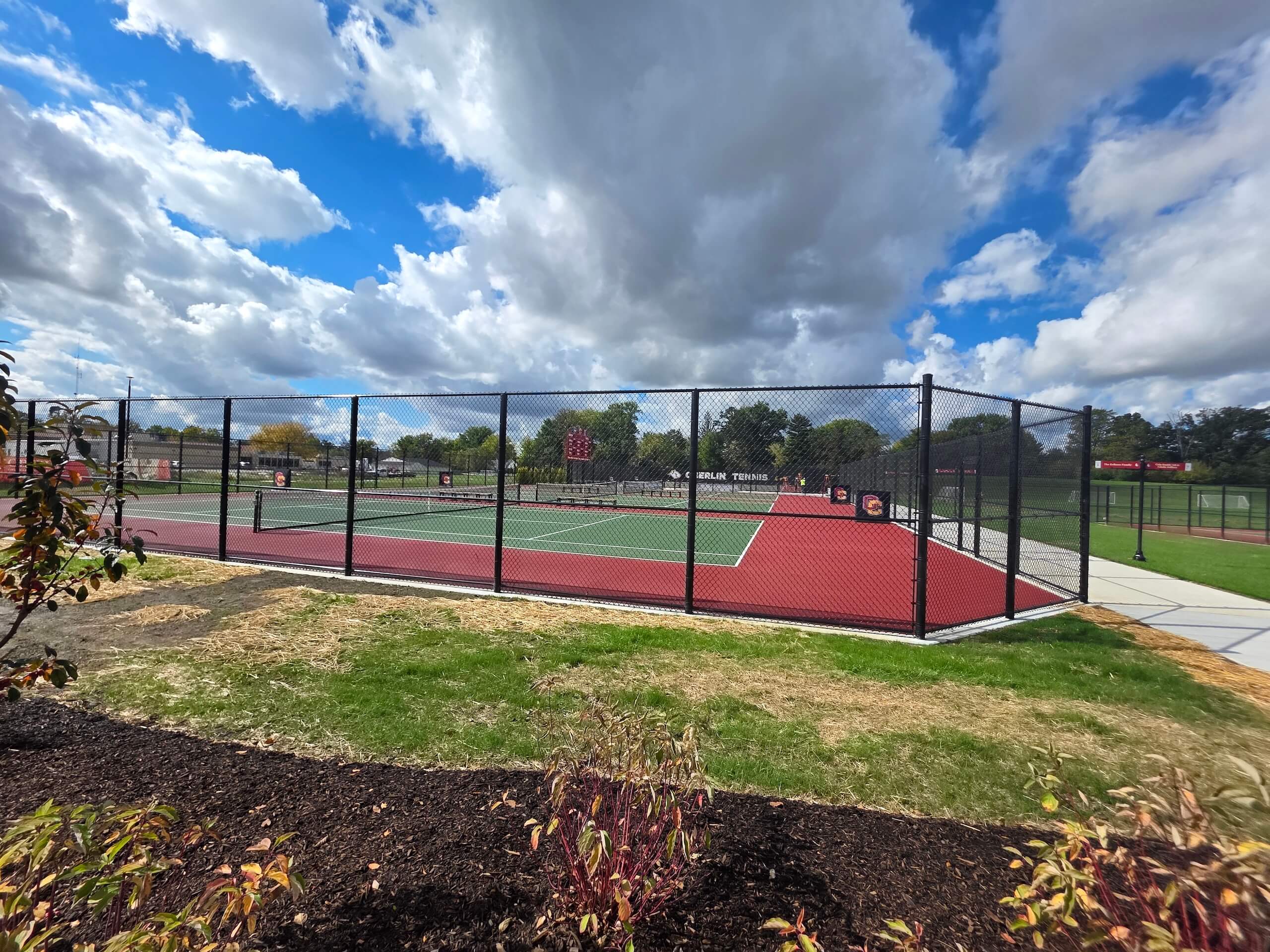 A fenced outdoor tennis court with red and green surfaces sits under a blue sky with scattered clouds, surrounded by grass, bushes, and a walking path.