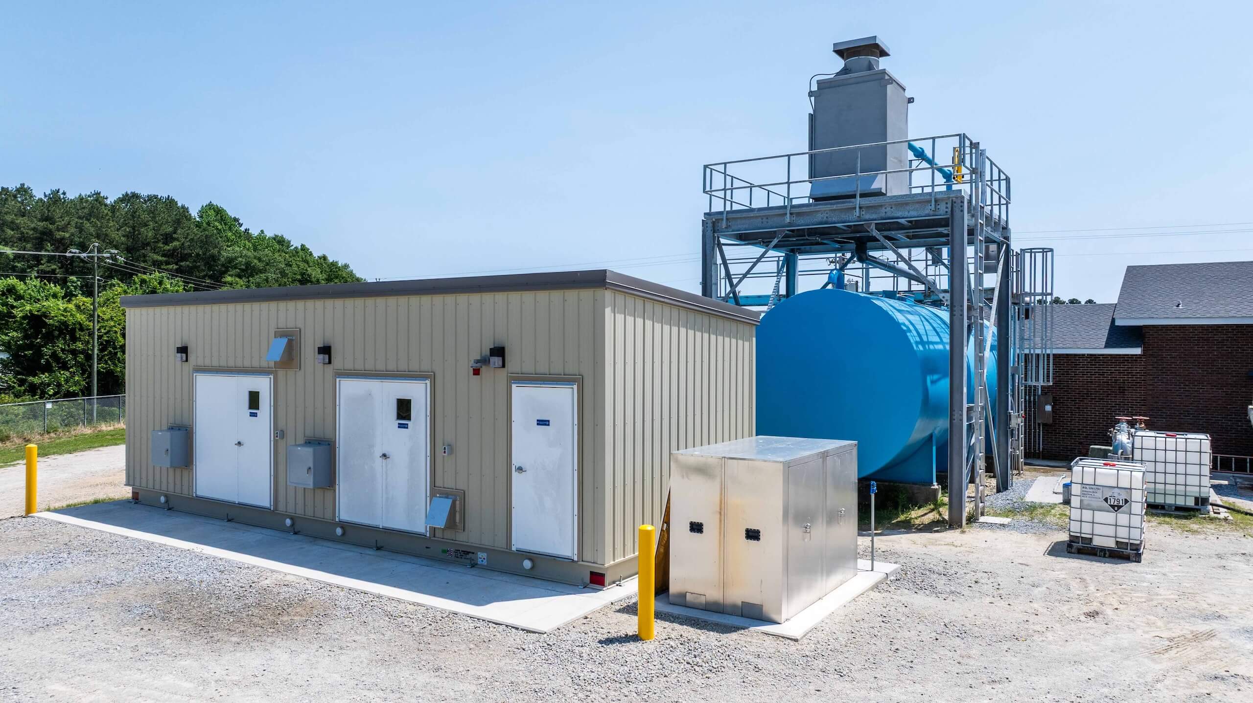 A small beige industrial building beside a large blue storage tank, metal structures, and various equipment, with trees and a clear sky in the background.