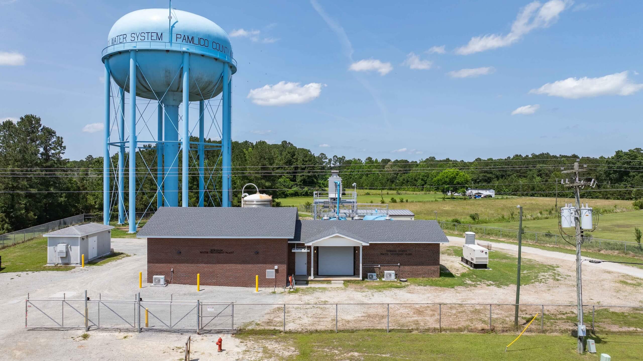 A tall blue water tower marked Water System, Pamlico County stands behind a brick utility building, fenced in and surrounded by grass, trees, and a partly cloudy sky.