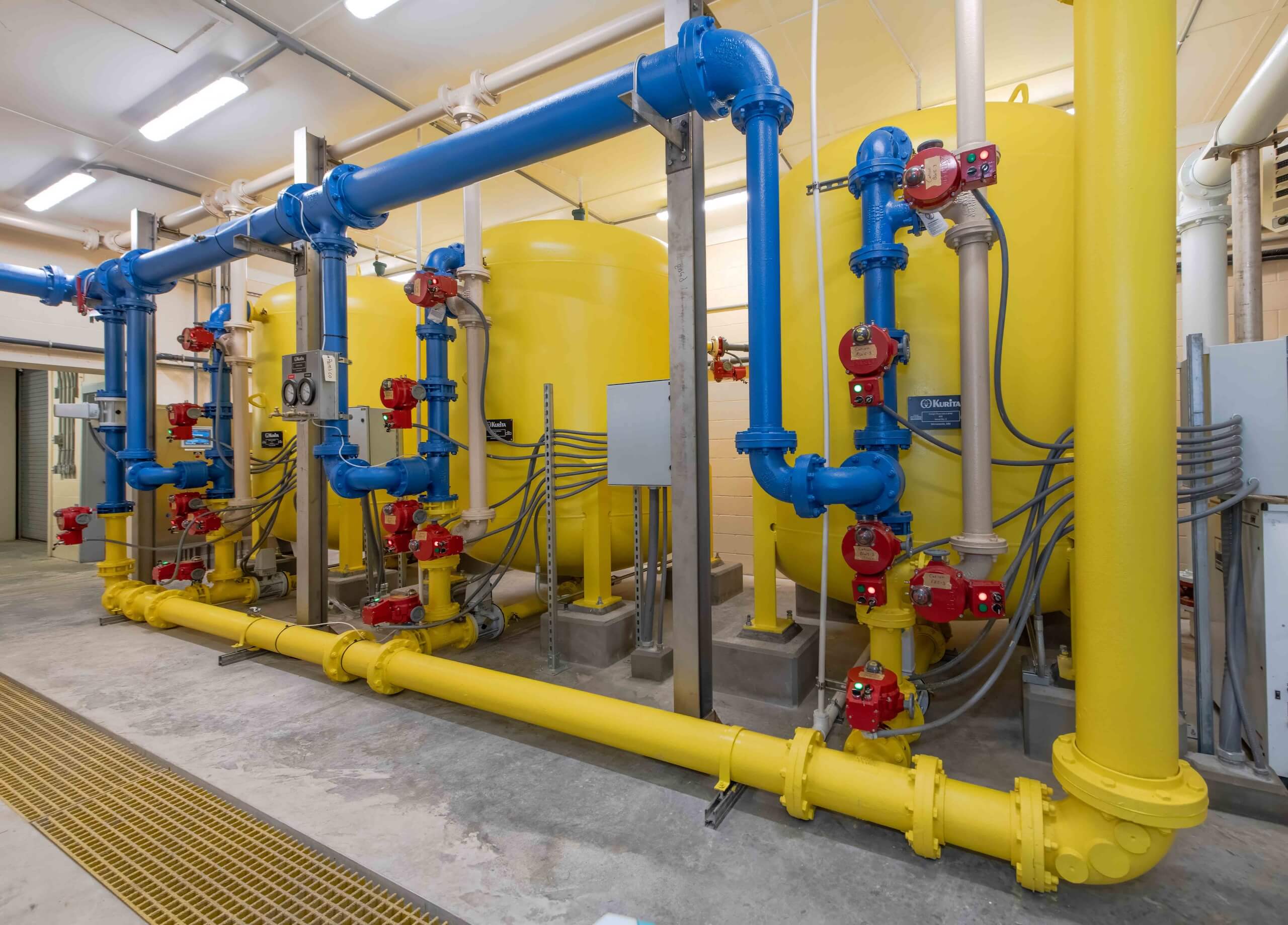Large yellow industrial tanks connected with blue and yellow pipes, featuring multiple red valves and gauges, inside a clean, well-lit facility with a concrete floor.