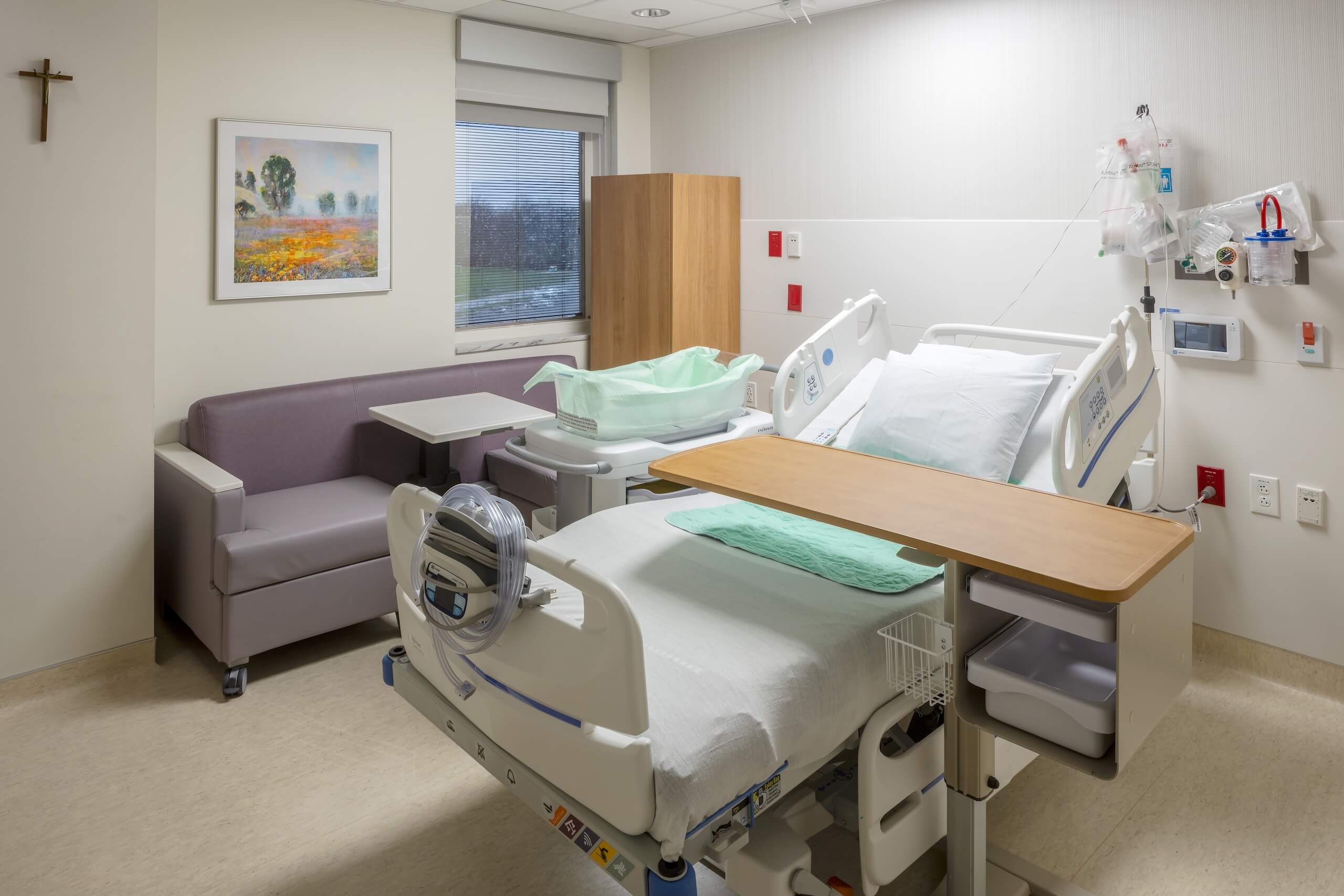A clean, modern hospital room with a single bed, a couch, a bassinet, medical equipment, a window, and artwork on the wall.