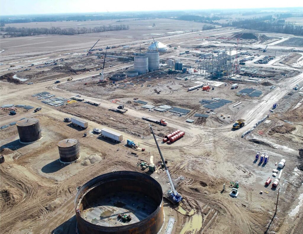 Aerial view of a large construction site with cranes, steel structures, industrial tanks, and scattered materials on muddy ground, surrounded by flat farmland under a cloudy sky.
