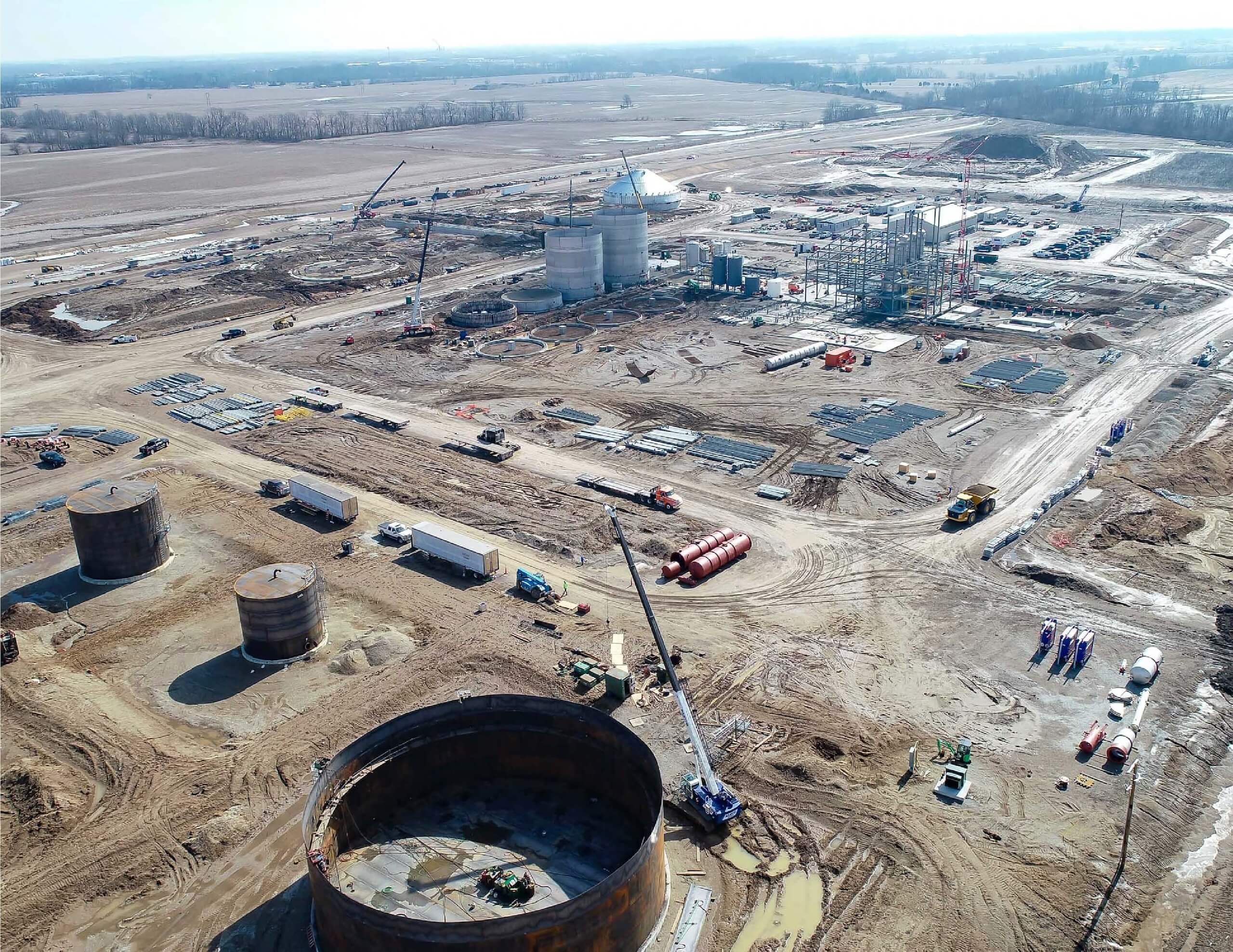 Aerial view of a large construction site with cranes, steel structures, industrial tanks, and scattered materials on muddy ground, surrounded by flat farmland under a cloudy sky.