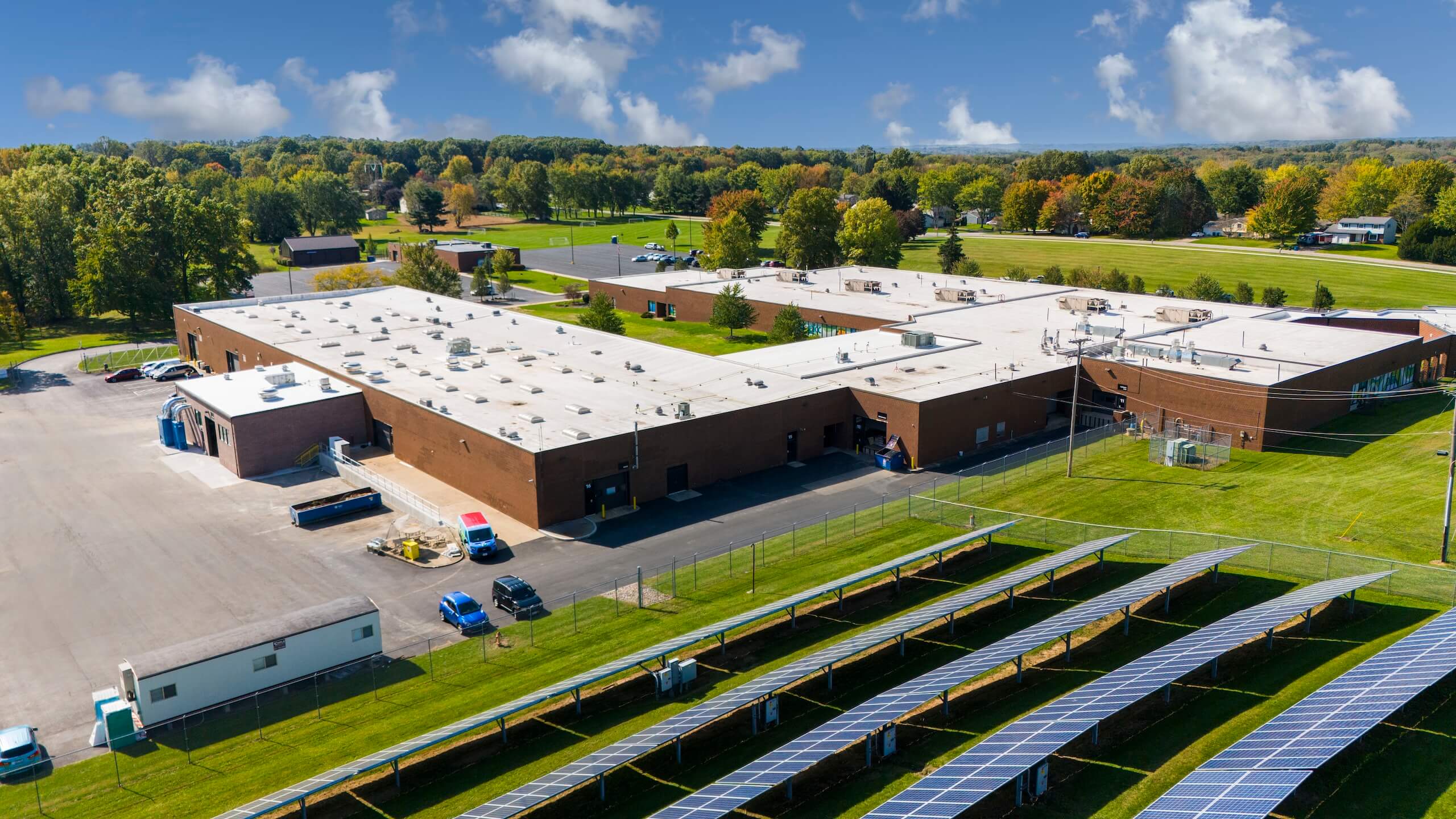 A large, flat-roofed brick building is surrounded by green lawns and trees. Rows of solar panels are visible in the foreground, with parked cars and a few small structures nearby. Blue sky with scattered clouds above.