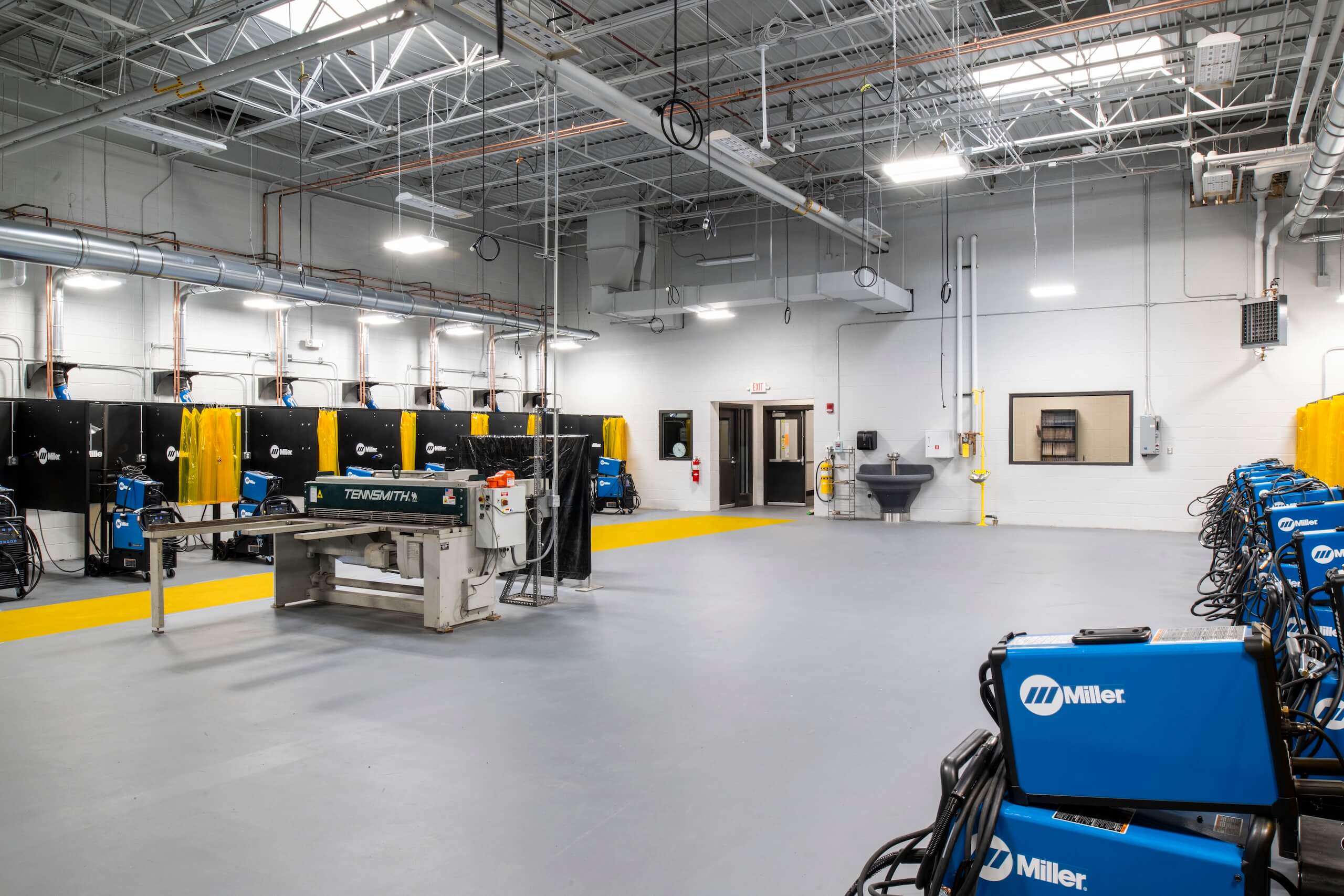 A spacious, clean industrial workshop with welding booths, blue Miller welding machines, yellow safety screens, metal tables, and exposed ceiling pipes under bright lighting.