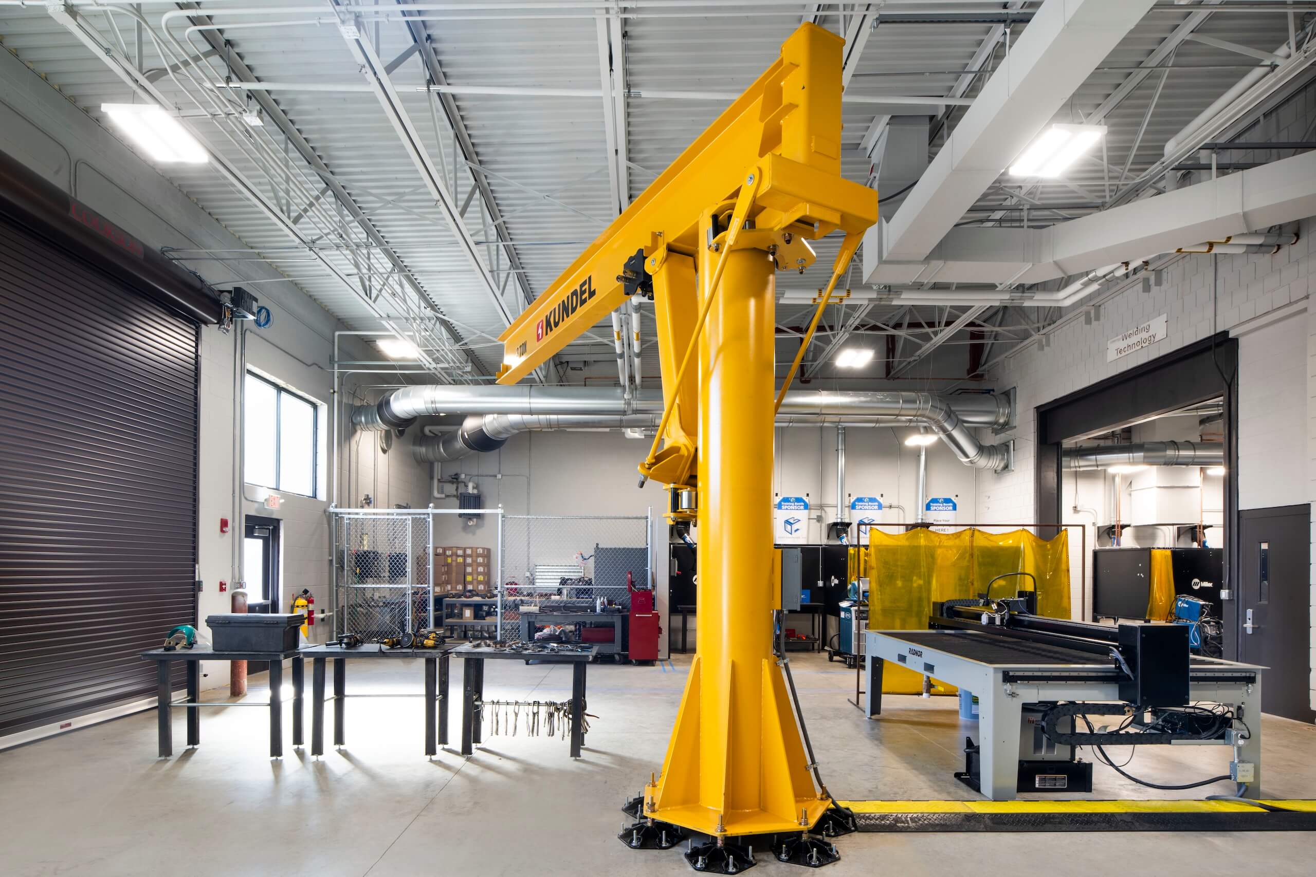 A bright yellow industrial jib crane stands in a spacious workshop with tables, equipment, metal fencing, and large ventilation ducts visible throughout the well-lit facility.