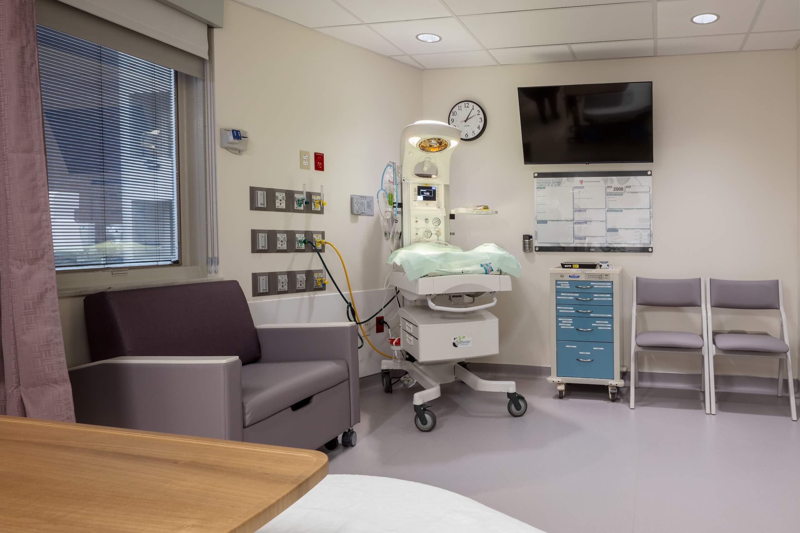 A modern hospital room with a baby warmer, medical equipment, a purple armchair, two white chairs, a wall TV, and a window with blinds. The environment is clean and organized.