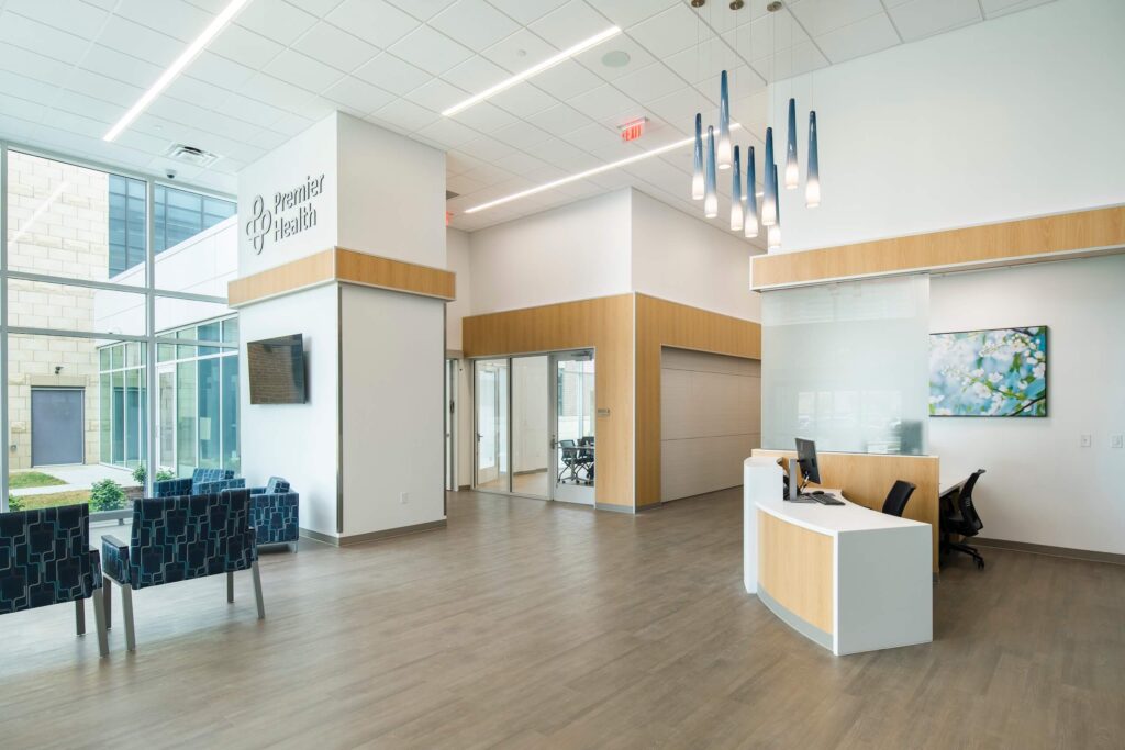Modern medical office lobby with a reception desk, blue chairs, large windows, light wood floors, and stylish hanging lights. Walls are white with some art and Premier Health signage. Bright, clean, and welcoming atmosphere.