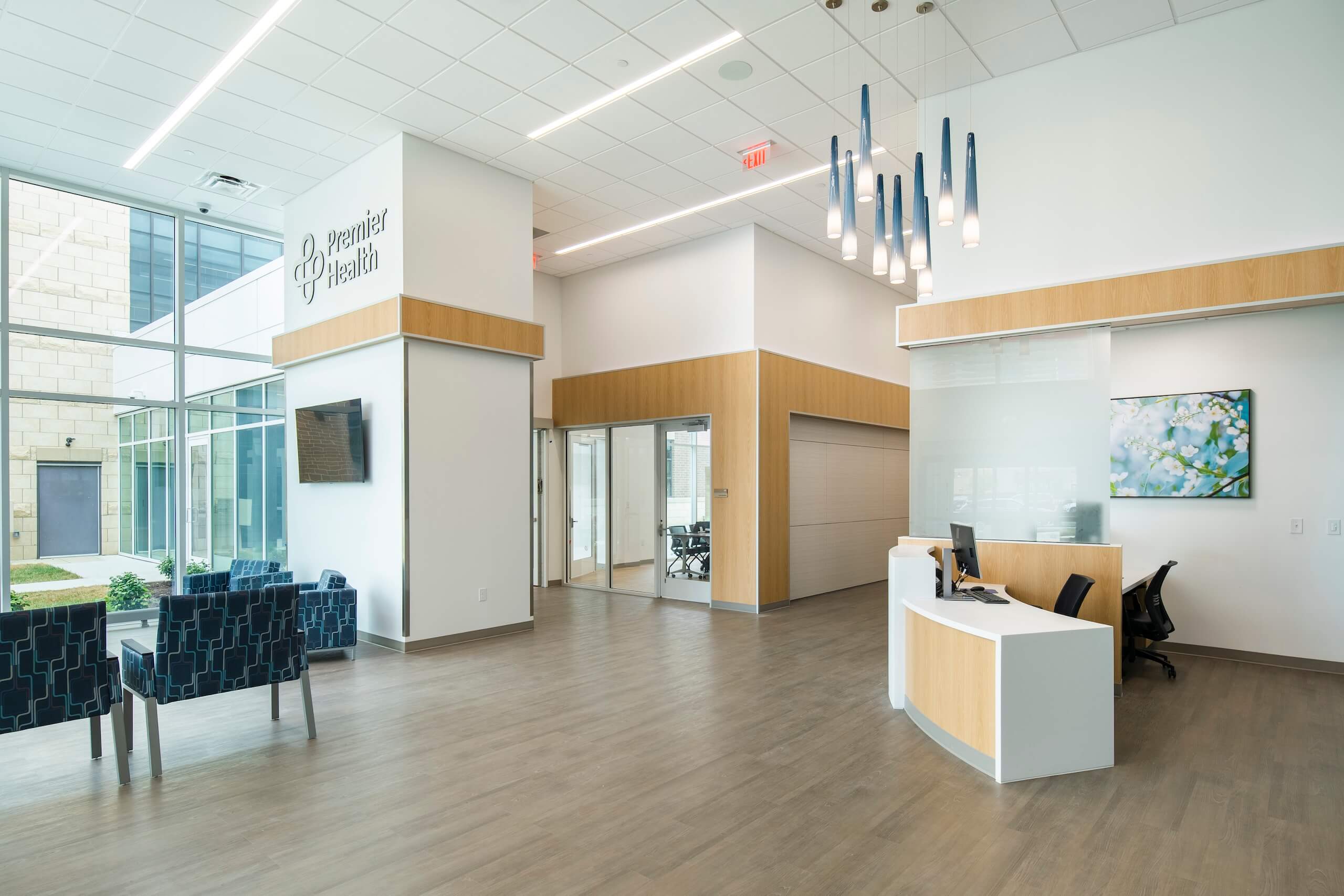 Modern medical office lobby with a reception desk, blue chairs, large windows, light wood floors, and stylish hanging lights. Walls are white with some art and Premier Health signage. Bright, clean, and welcoming atmosphere.
