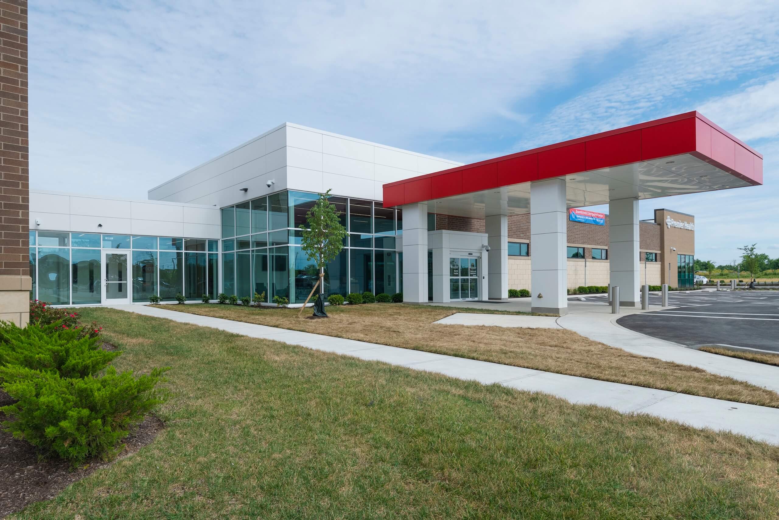 Modern building with large glass windows, a red canopy over the entrance, landscaped greenery, and a parking lot in front. The sky is partly cloudy.