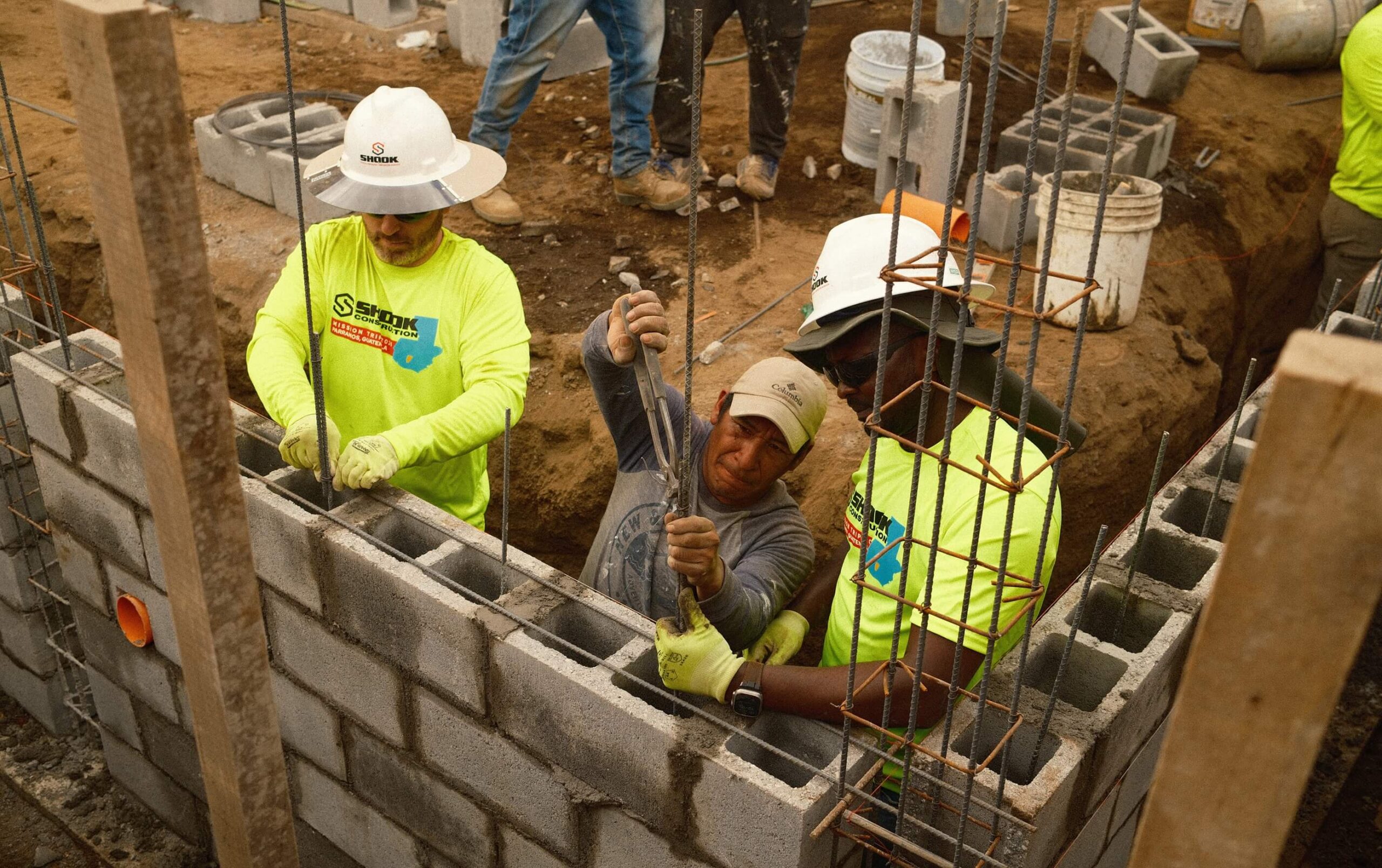 Three construction workers build a concrete block wall, inserting rebar and wearing safety gear and gloves. Dirt, cinder blocks, and construction tools are visible around the partially built wall.