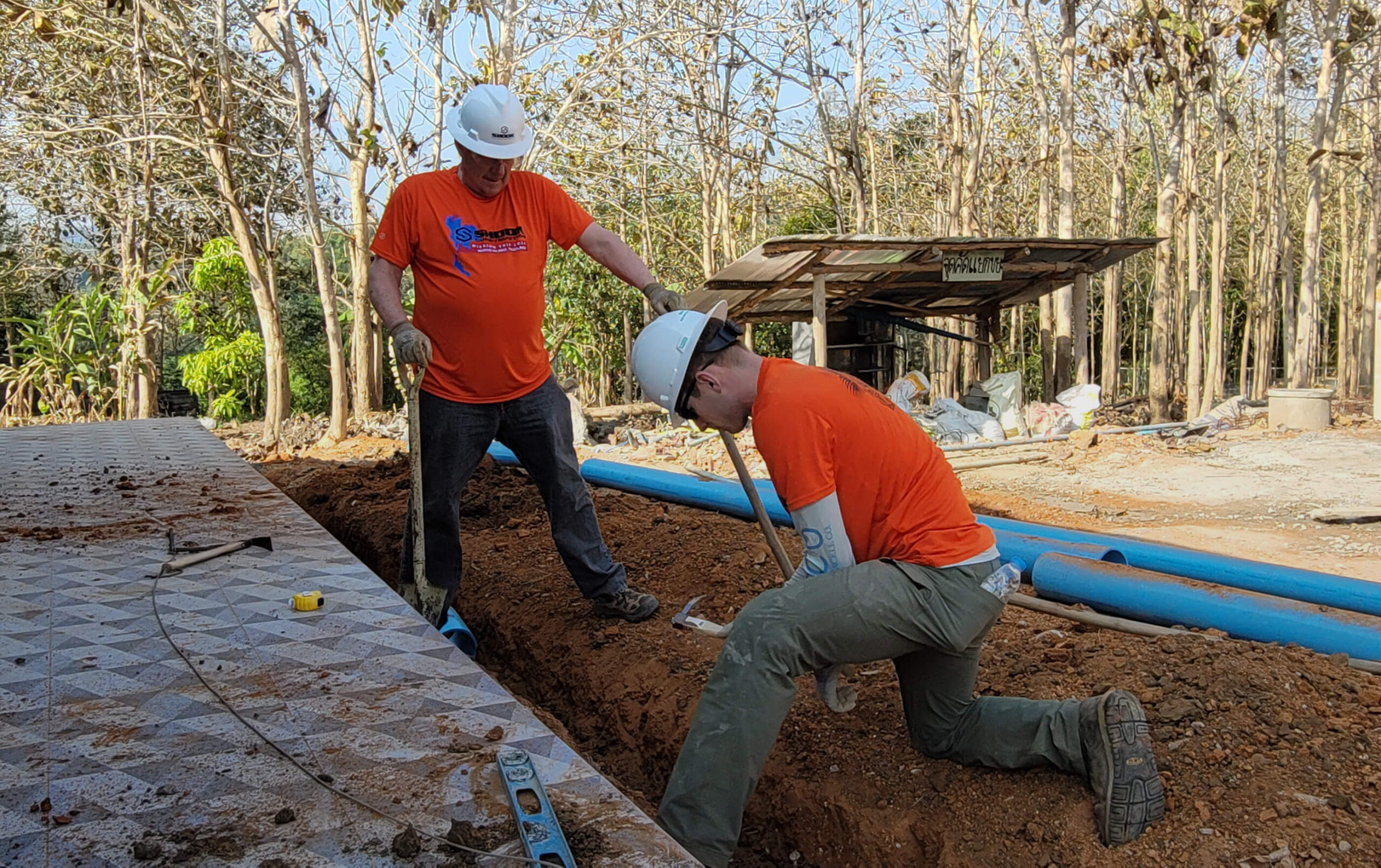 Two workers in orange shirts and white hard hats dig a trench near a tiled area outdoors, surrounded by trees and construction tools like a level and blue pipes.