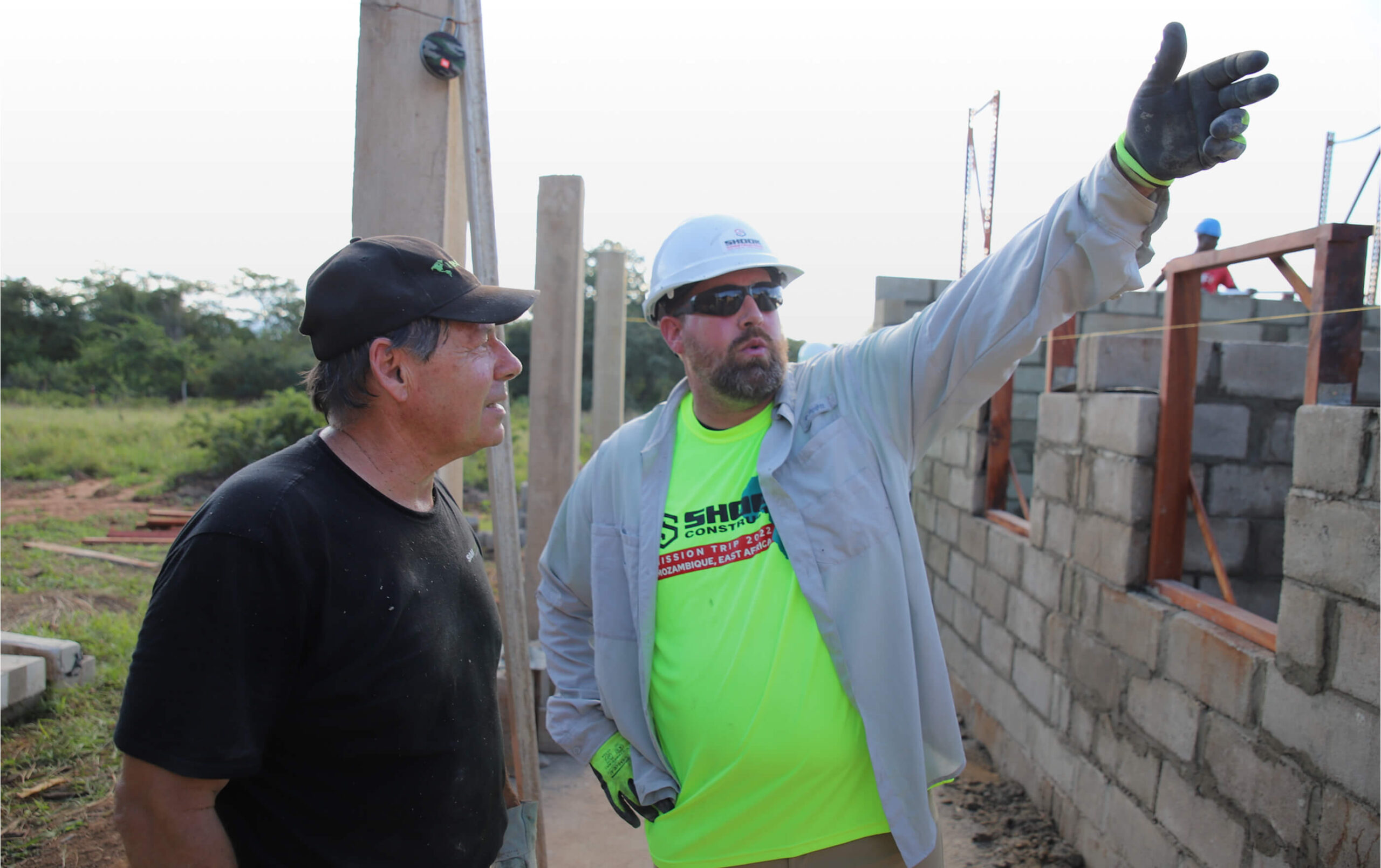Two construction workers stand at a building site. One in a neon shirt and hard hat points and gives directions, while the other listens attentively. They are surrounded by partially built concrete block walls.