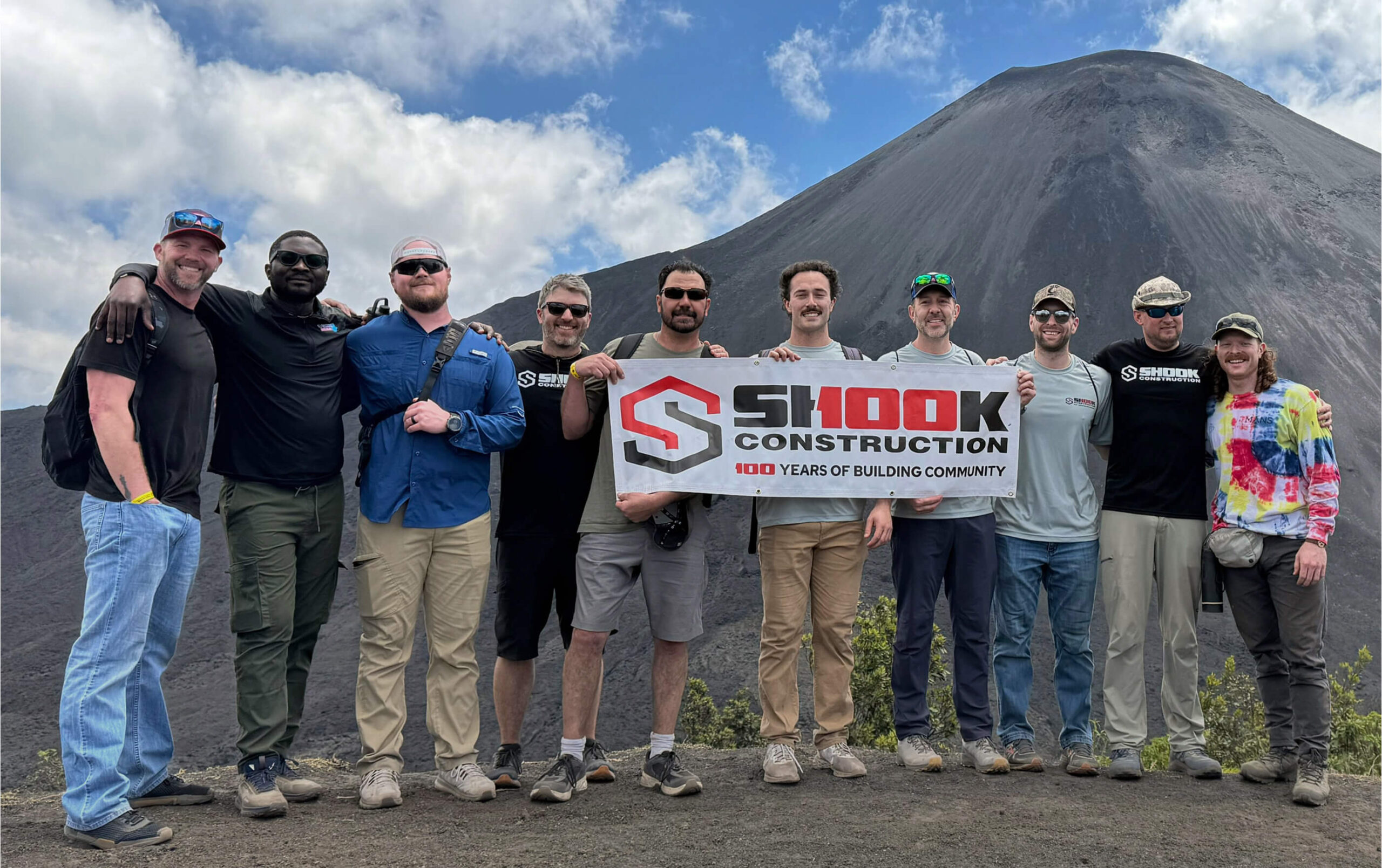 Ten people stand outdoors in front of a volcano, smiling and holding a Shook Construction: 100 Years of Building Community banner. The sky is partly cloudy, and everyone is dressed in casual outdoor clothing.