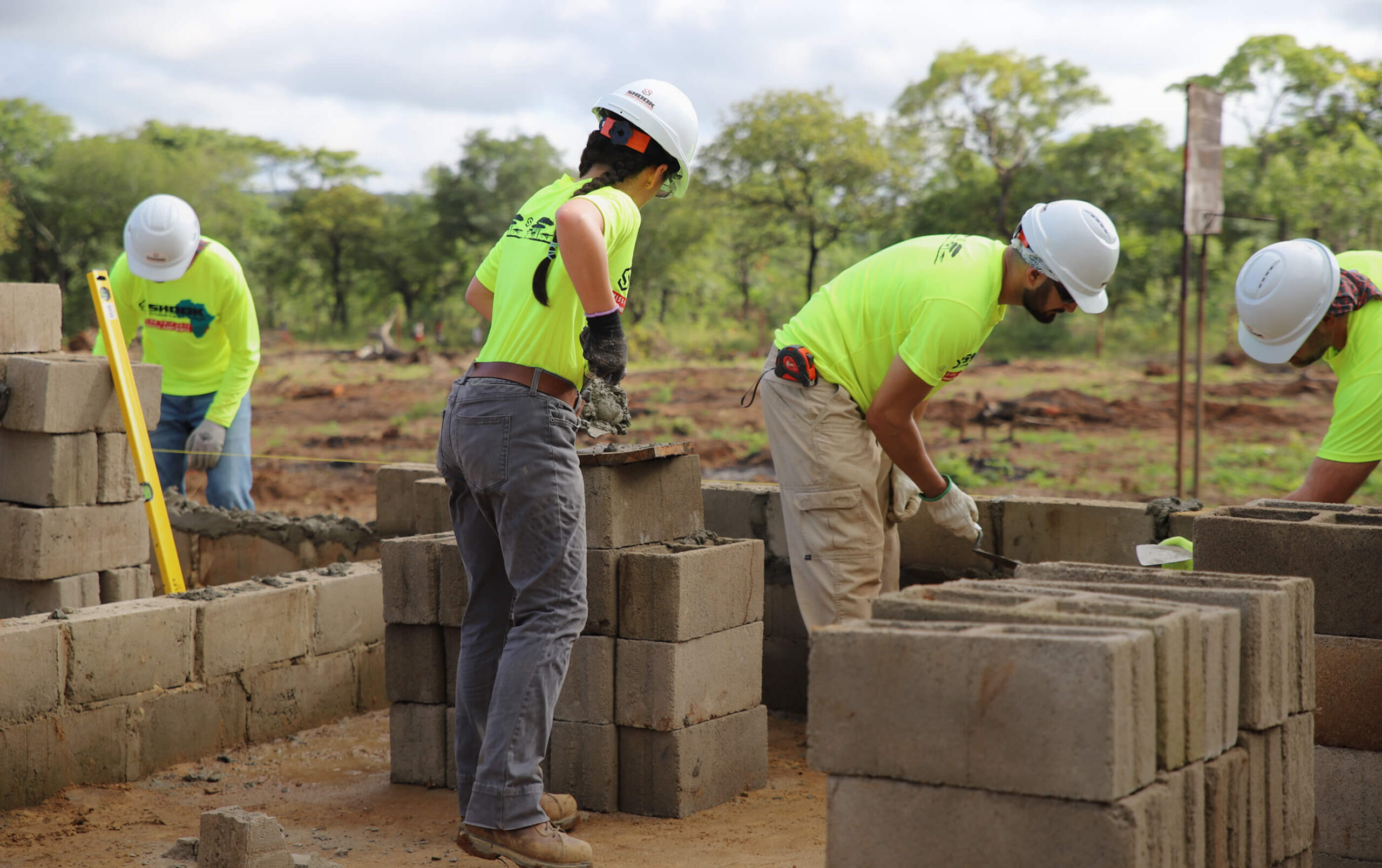 Four people wearing safety gear and neon shirts build a wall with concrete blocks outdoors on a construction site surrounded by greenery and trees.