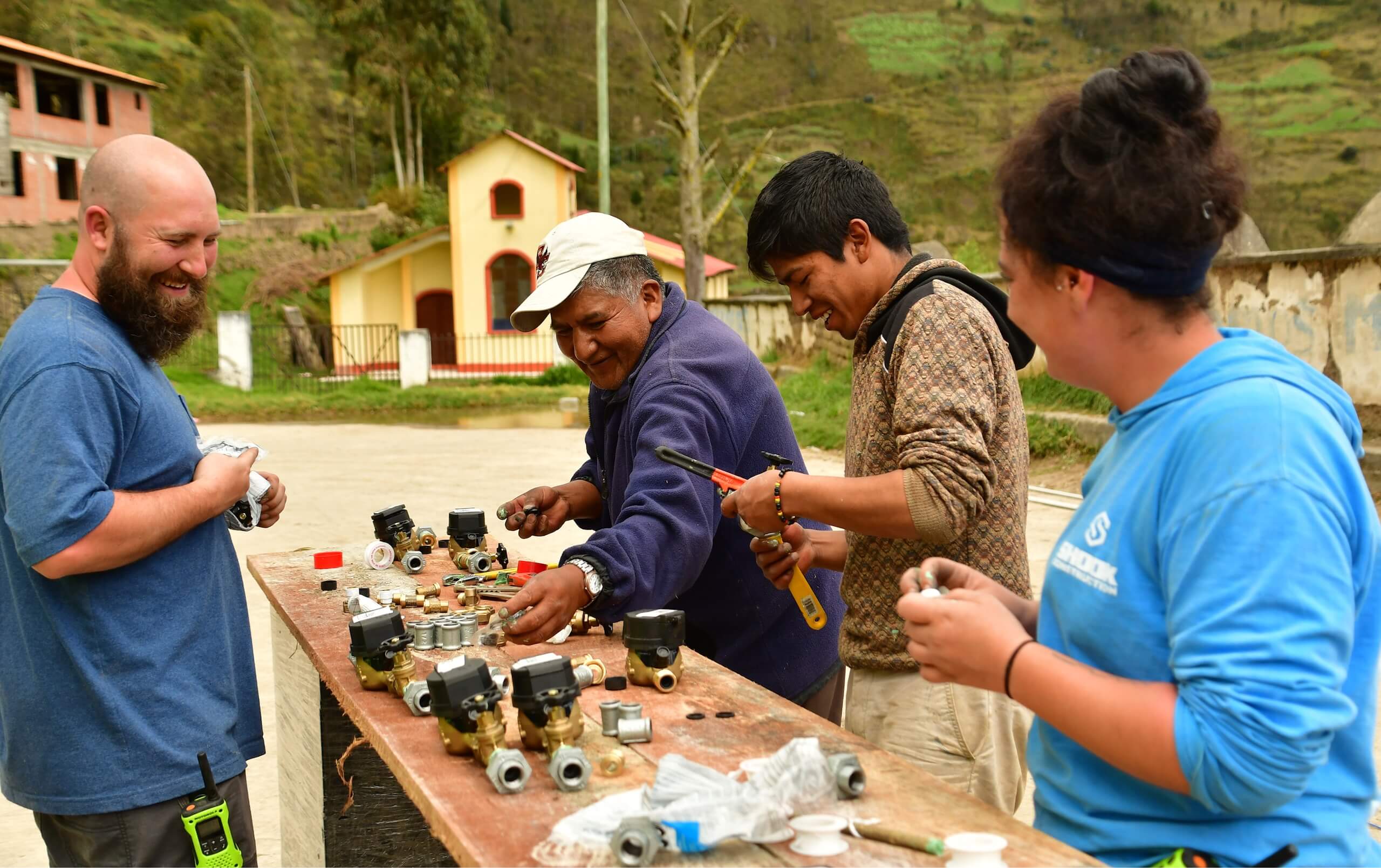 Four people work together outdoors at a wooden table assembling plumbing or mechanical parts. They are smiling and appear to be collaborating. Green hills and buildings are visible in the background.