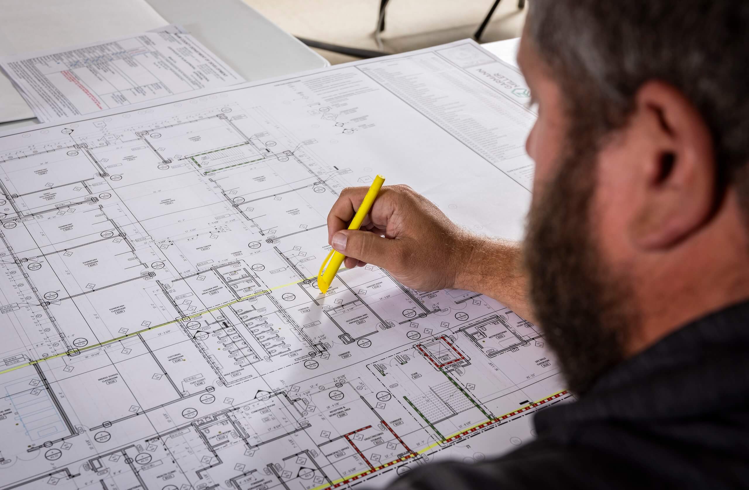 A person reviews a large architectural blueprint, using a yellow highlighter to mark sections on the detailed floor plan spread out on a table.