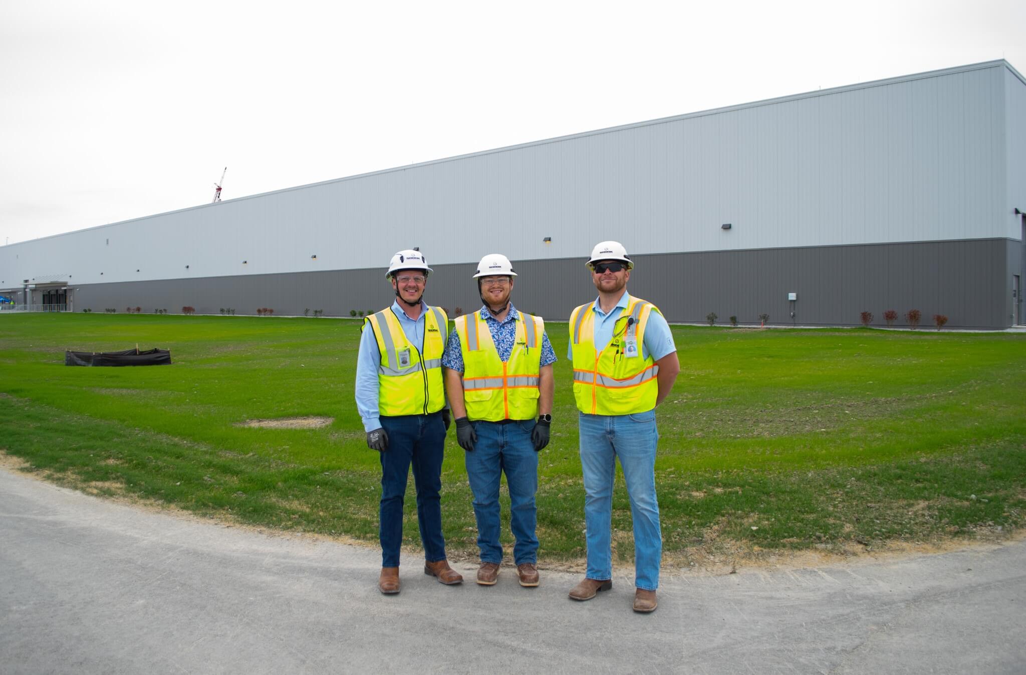 Three construction workers in safety vests, hard hats, and gloves stand smiling in front of a large industrial building on a grassy area.