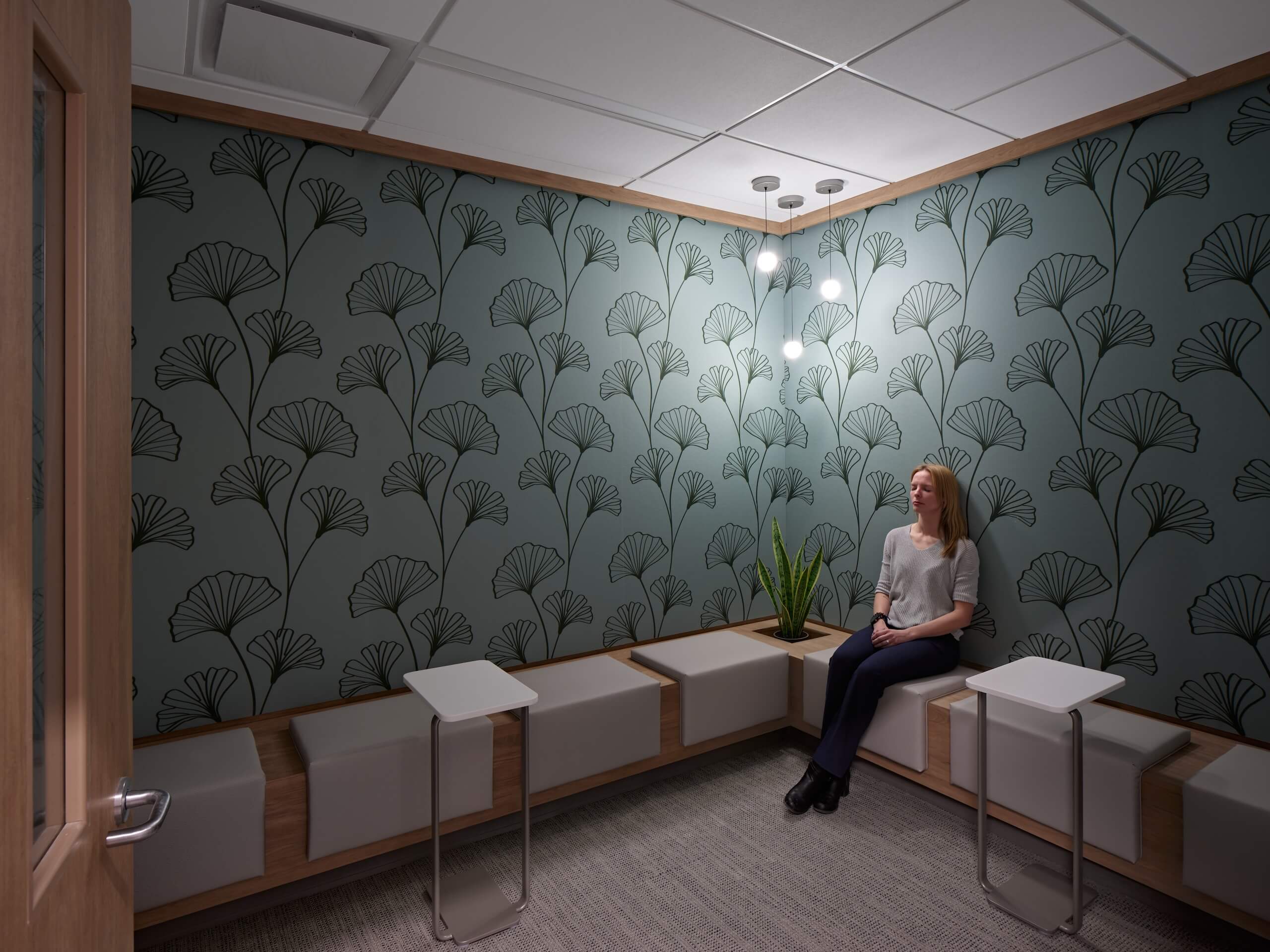 A woman sits alone on a bench in a small modern room with patterned wallpaper, soft lighting, and several small tables and a potted plant.