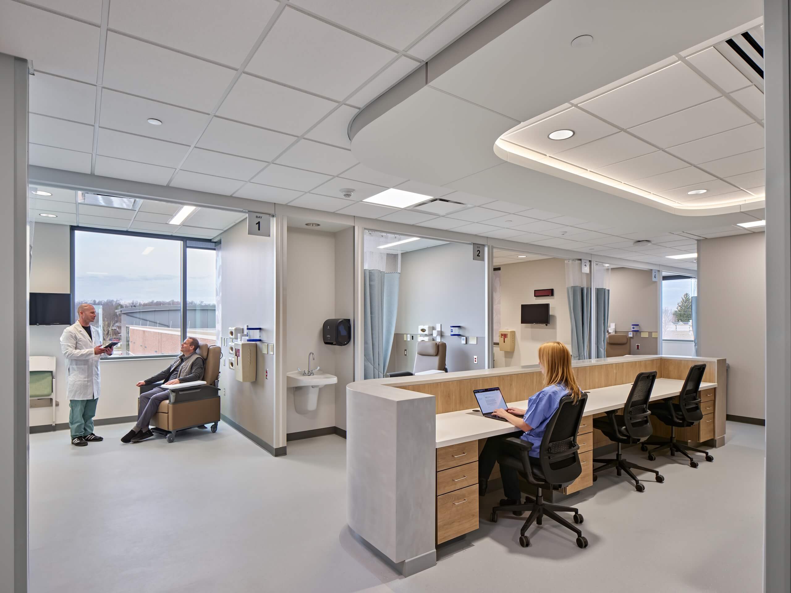 A modern hospital room with a nurse working at a computer, a doctor talking to a patient in a chair, and medical equipment visible. The space is clean, bright, and organized.