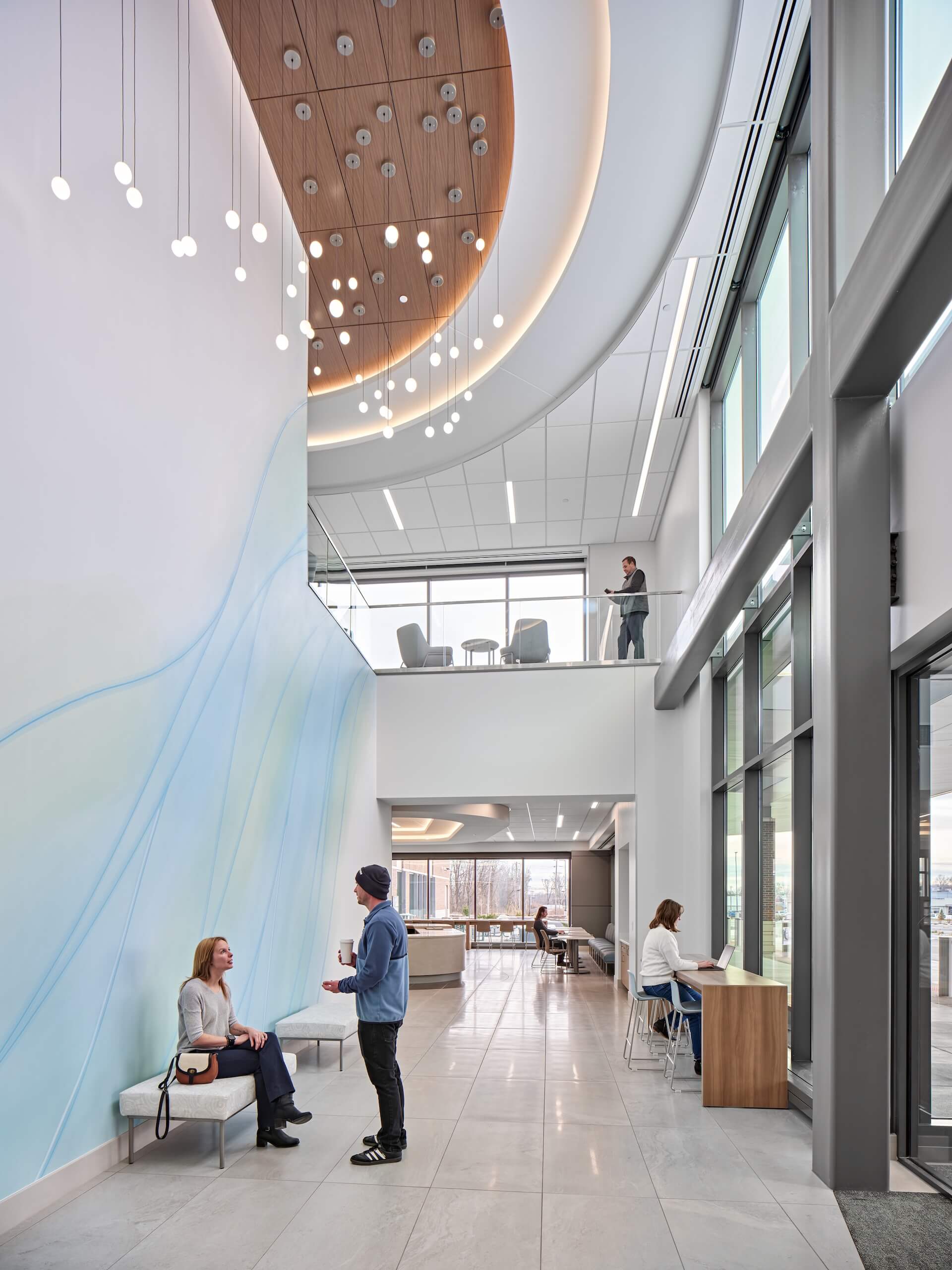 Modern lobby with high ceilings, large windows, hanging lights, and a blue accent wall. People are sitting, talking, and working on two levels in a bright, inviting space.