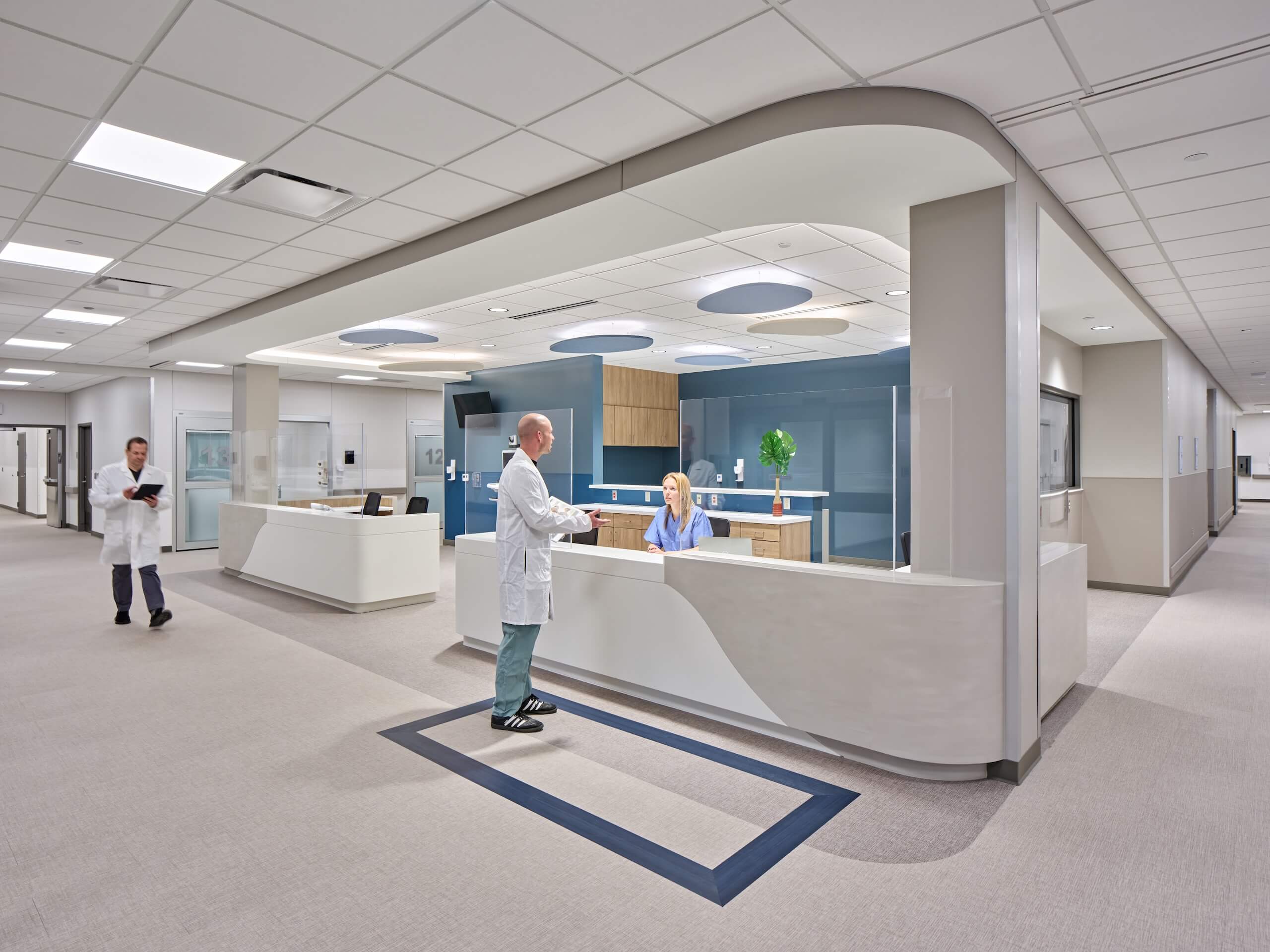 Modern hospital reception area with two medical staff talking at the front desk and another staff member walking down a wide, well-lit hallway. The space is clean, bright, and minimally decorated.