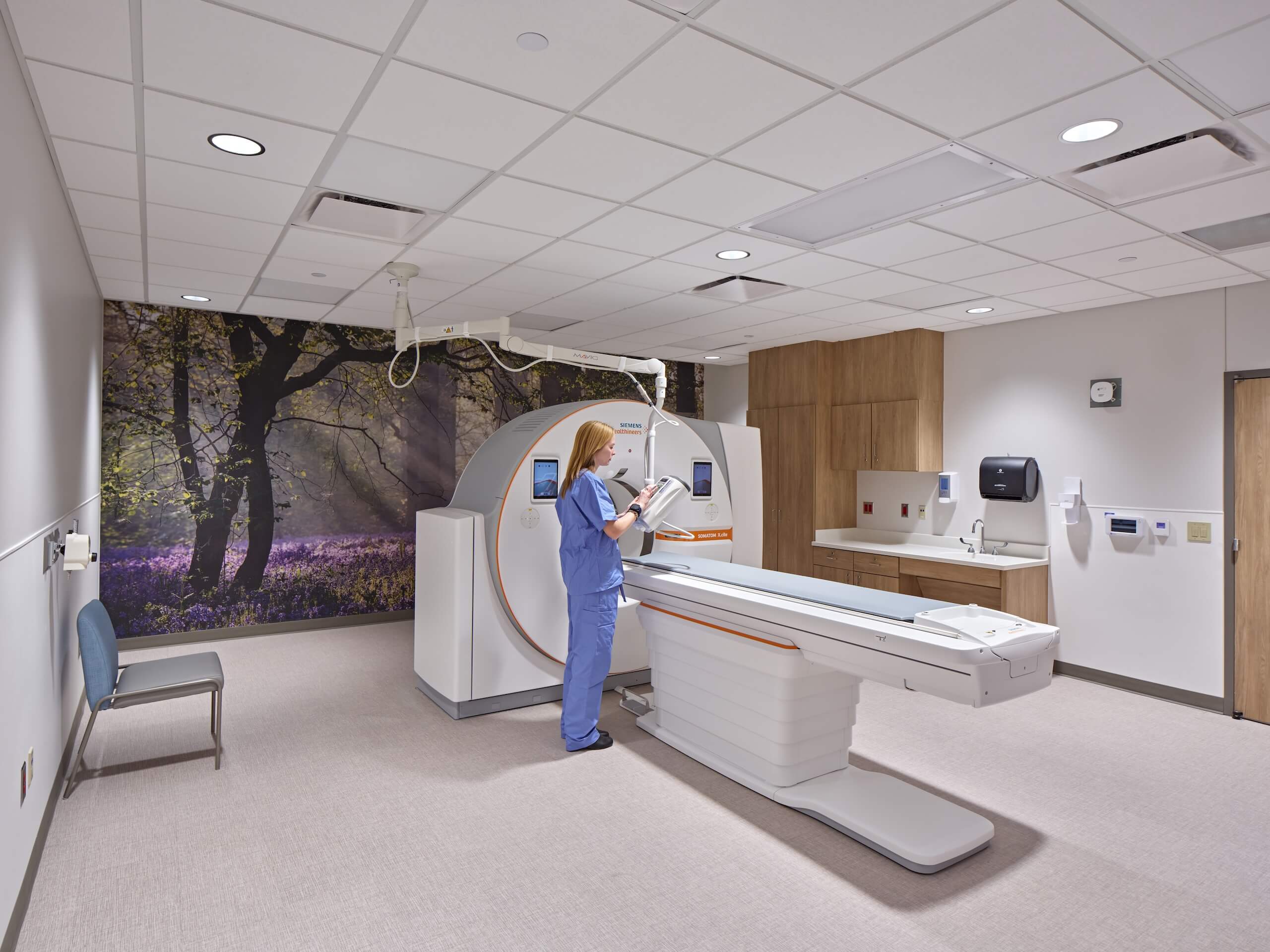 A medical professional in scrubs stands by a CT scanner in a clean, modern imaging room with woodland wall art, checking a monitor or clipboard.