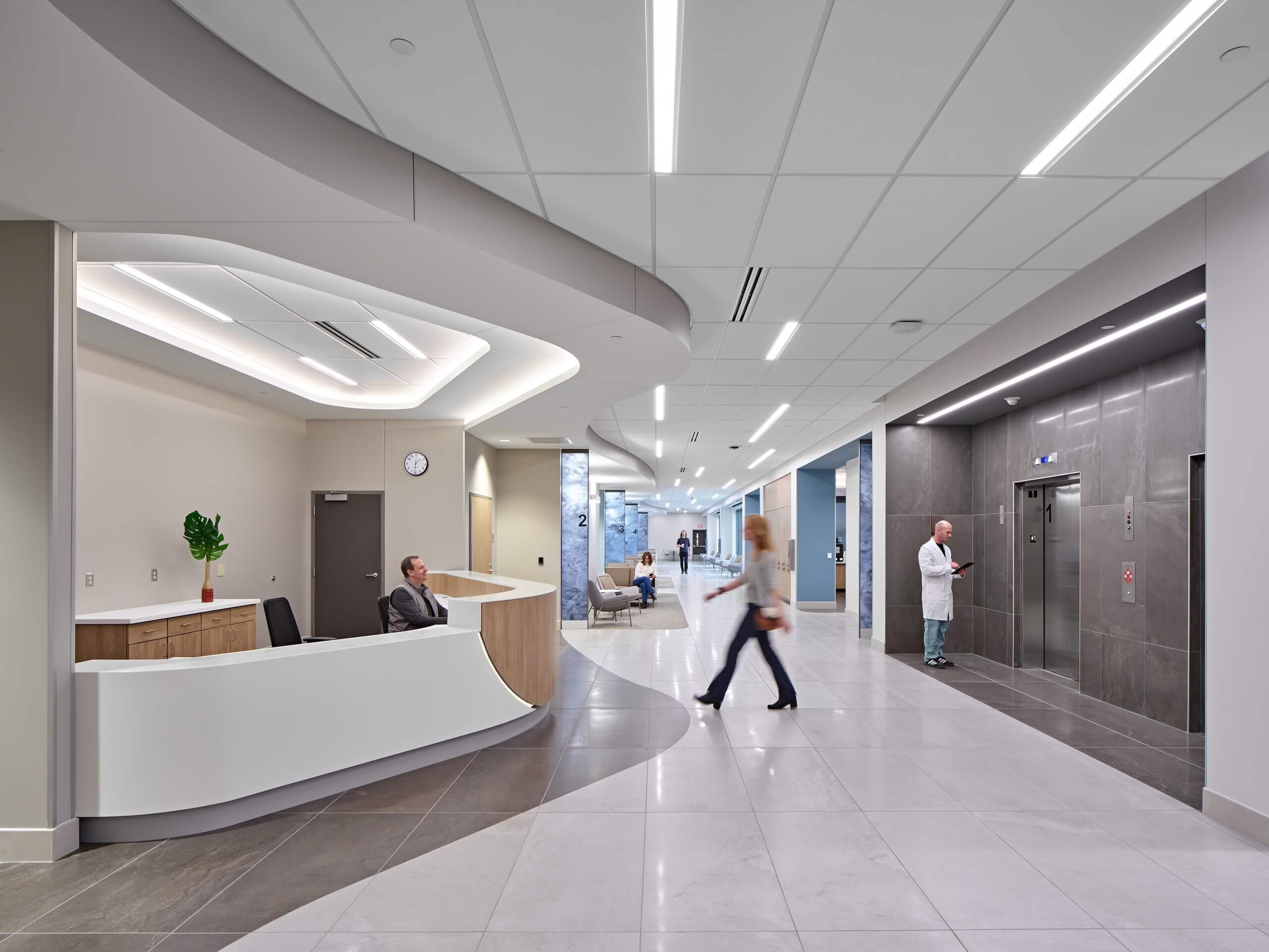 A modern hospital lobby with a curved reception desk, a seated receptionist, a person walking, a doctor waiting by elevators, and bright overhead lighting.
