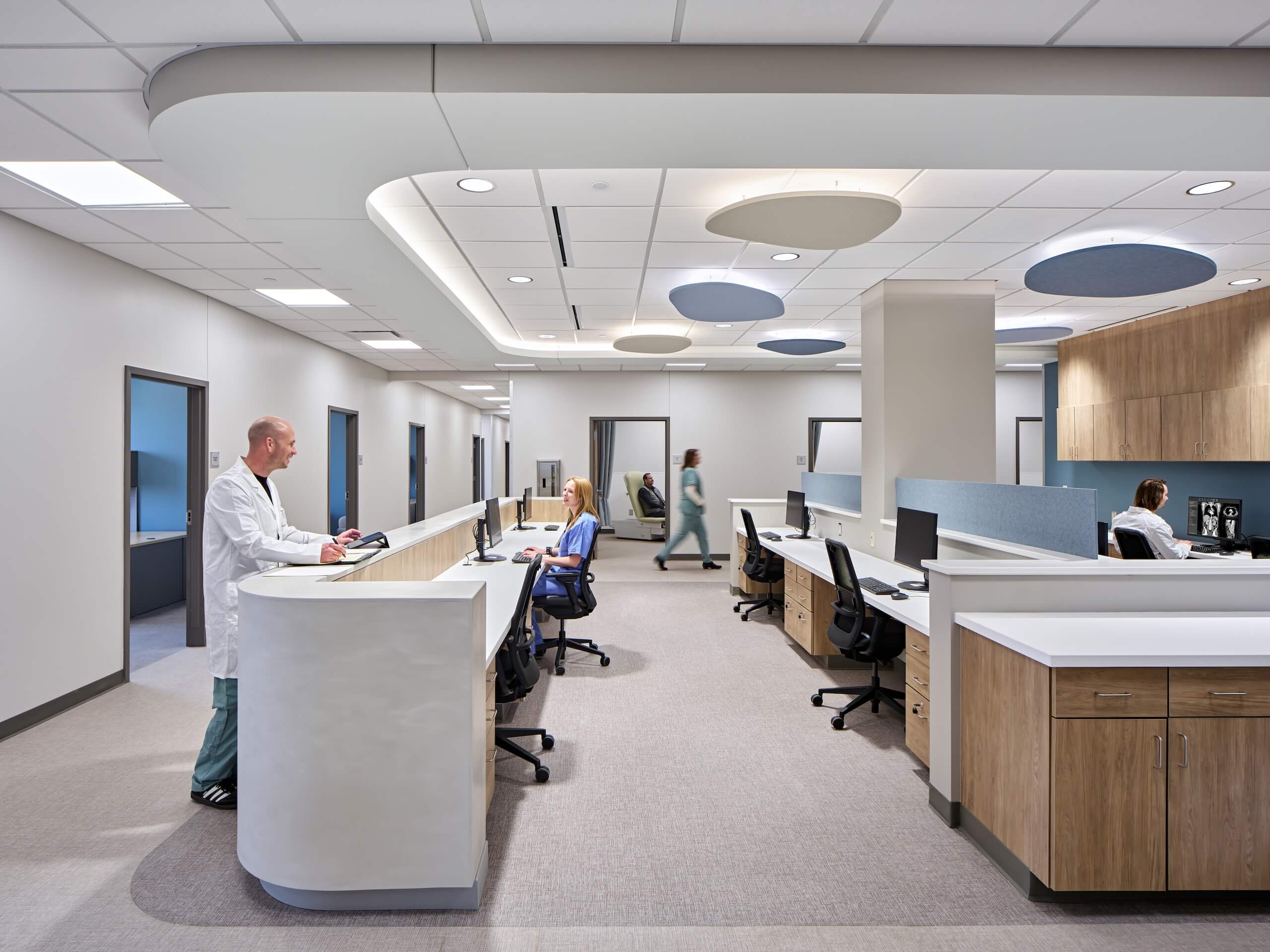 Modern medical office with a doctor at a curved reception desk, a nurse seated at a computer, and other staff walking in the background. The space is clean, bright, and organized with wood cabinetry and white countertops.