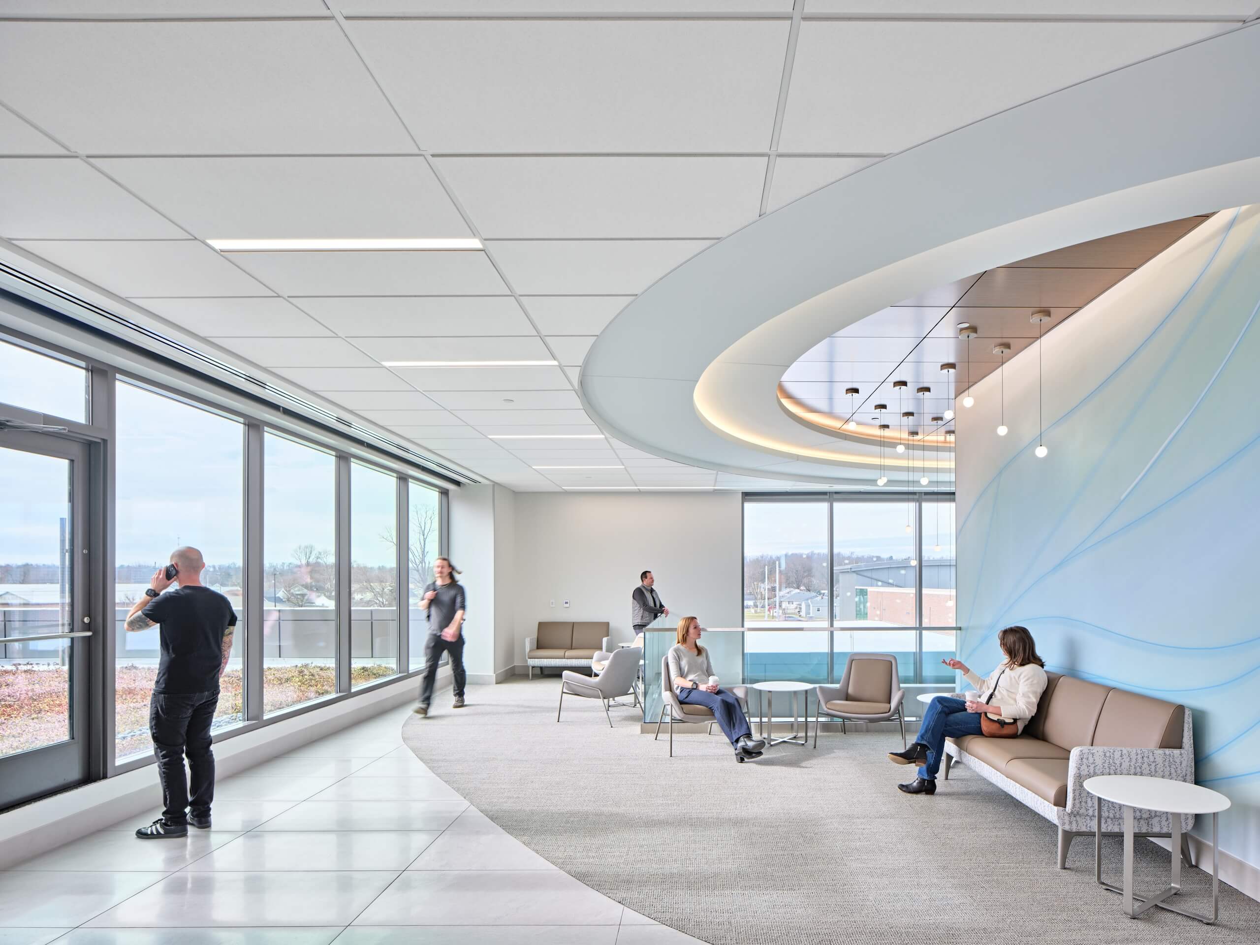Modern office lounge with large windows, curved ceiling details, and several people sitting or standing. Some are talking, one is on the phone, and one is walking. Neutral colors dominate the space, creating a calm atmosphere.
