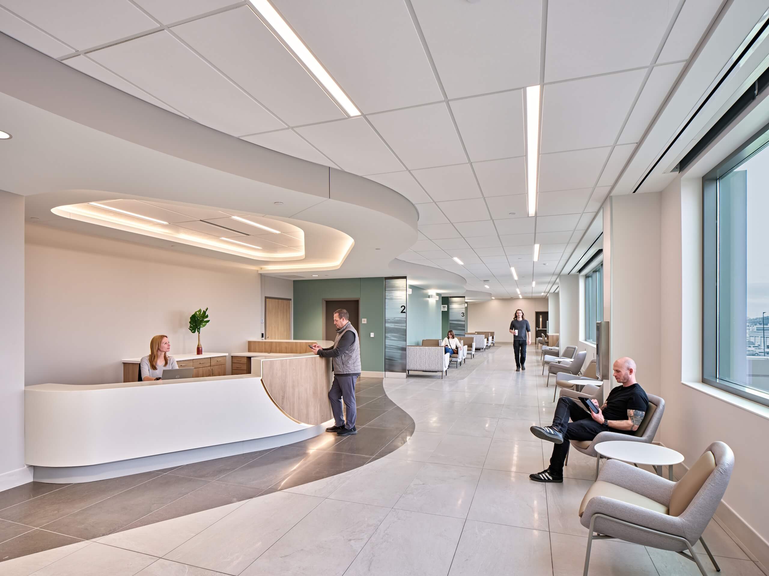 A modern medical office waiting area with a receptionist at a desk, a man talking to her, several people seated, and large windows letting in natural light.