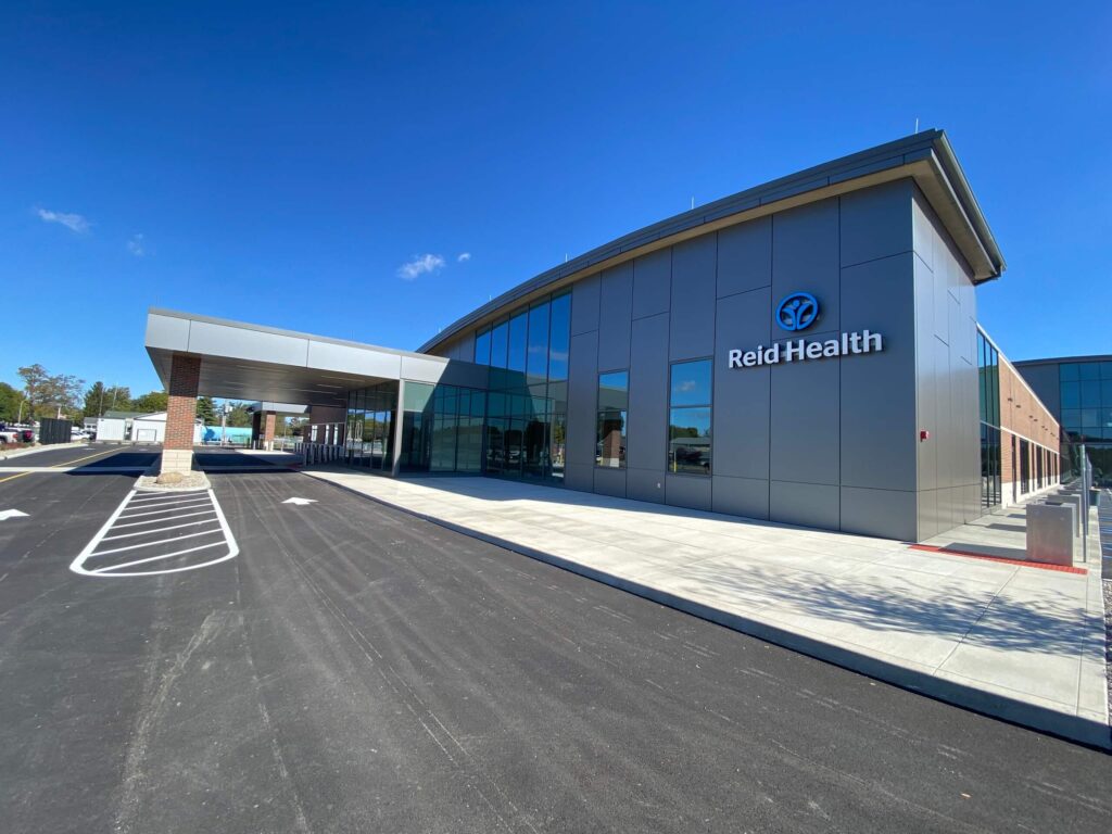 A modern building with large windows and a sign that reads Reid Health is shown under a clear blue sky. The entrance has a covered drop-off area and an empty parking lot in front.