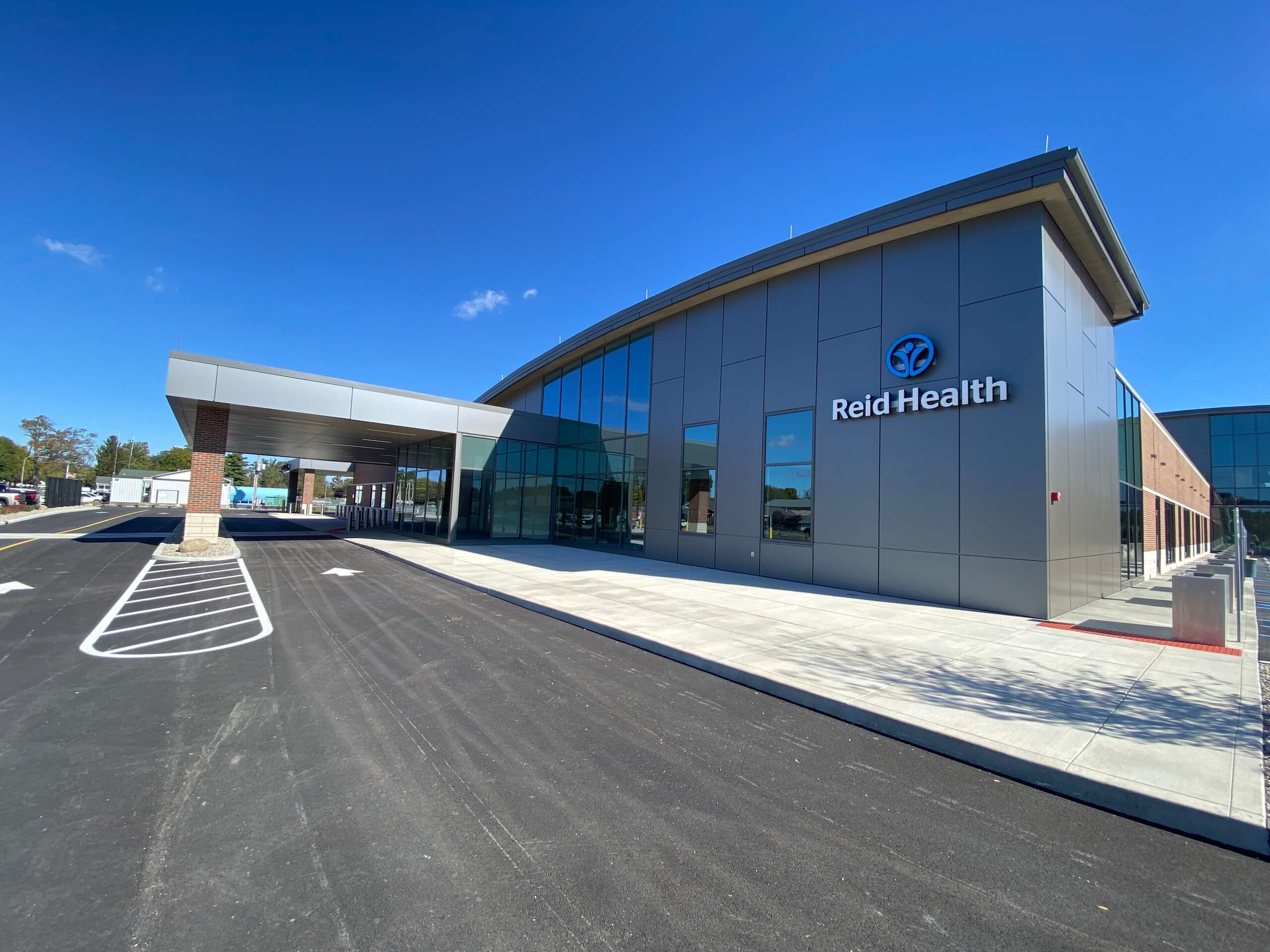A modern building with large windows and a sign that reads Reid Health is shown under a clear blue sky. The entrance has a covered drop-off area and an empty parking lot in front.