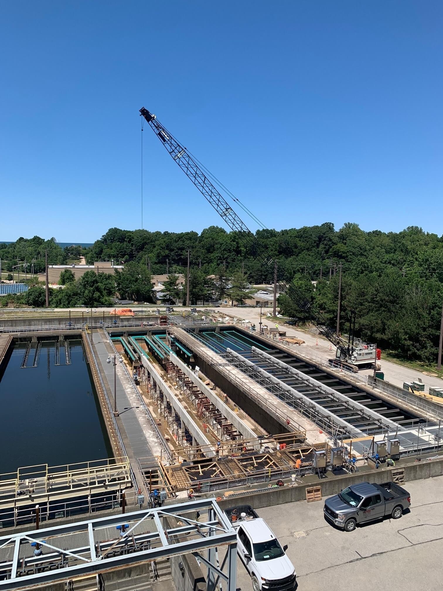 A water treatment facility under construction, with a crane, vehicles, and equipment on site. Trees and clear blue sky are visible in the background.