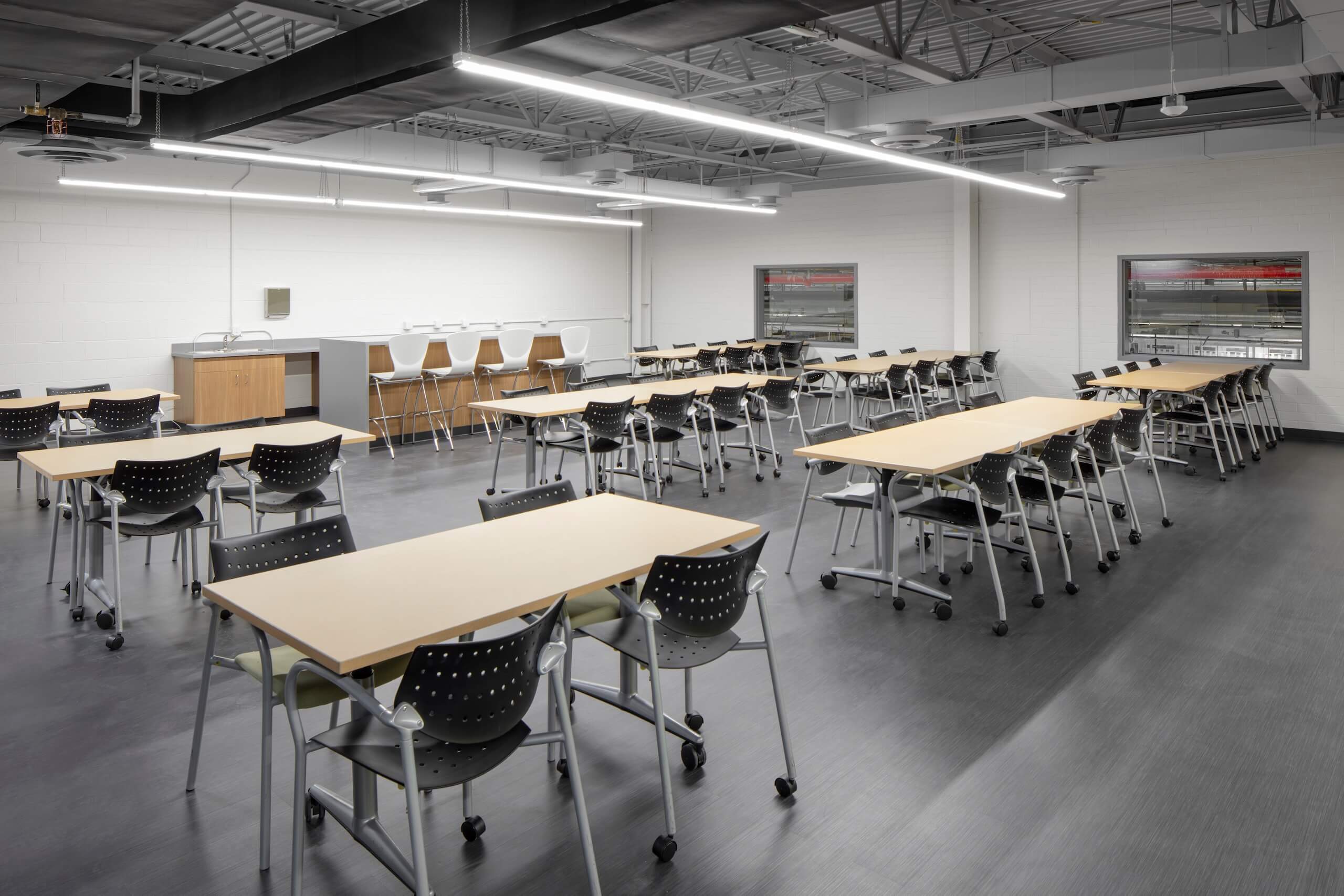 A modern classroom with several rectangular tables and black chairs on wheels, arranged in rows. There are tall stools and a counter along the back wall. The room has gray floors and bright overhead lighting.