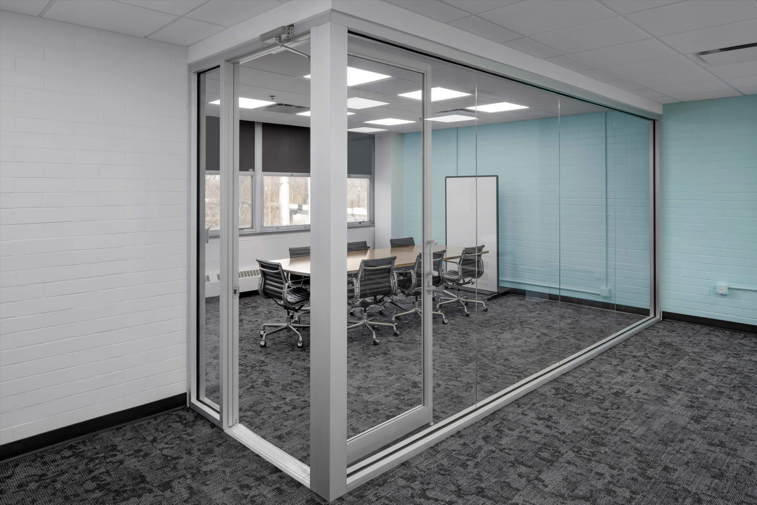 A modern glass-walled conference room with a table, eight chairs, a whiteboard, and windows letting in natural light. The walls are light blue and white, and the carpet is dark gray.