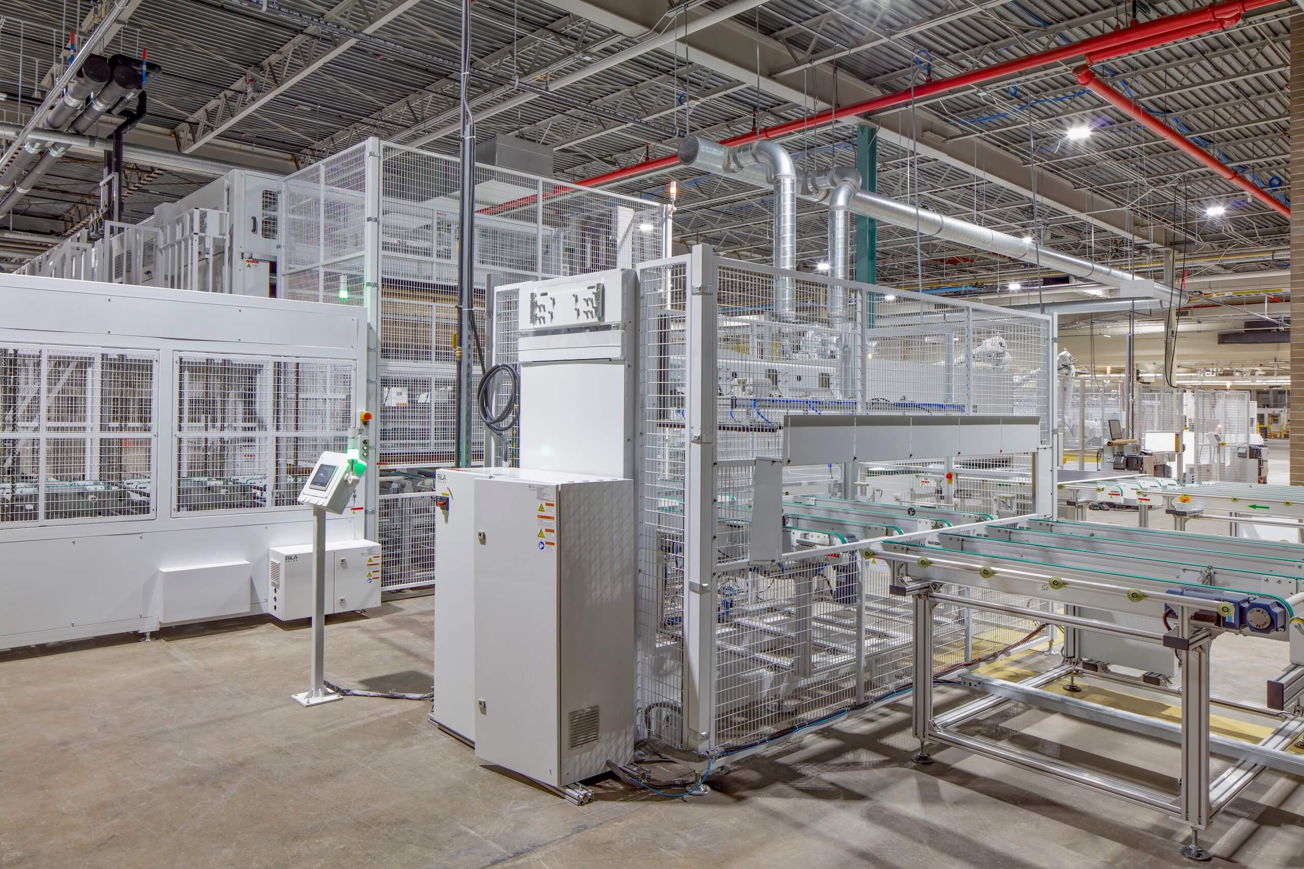 A modern factory floor with automated machinery, conveyor belts, and safety enclosures, under bright industrial lighting and exposed ceiling pipes.