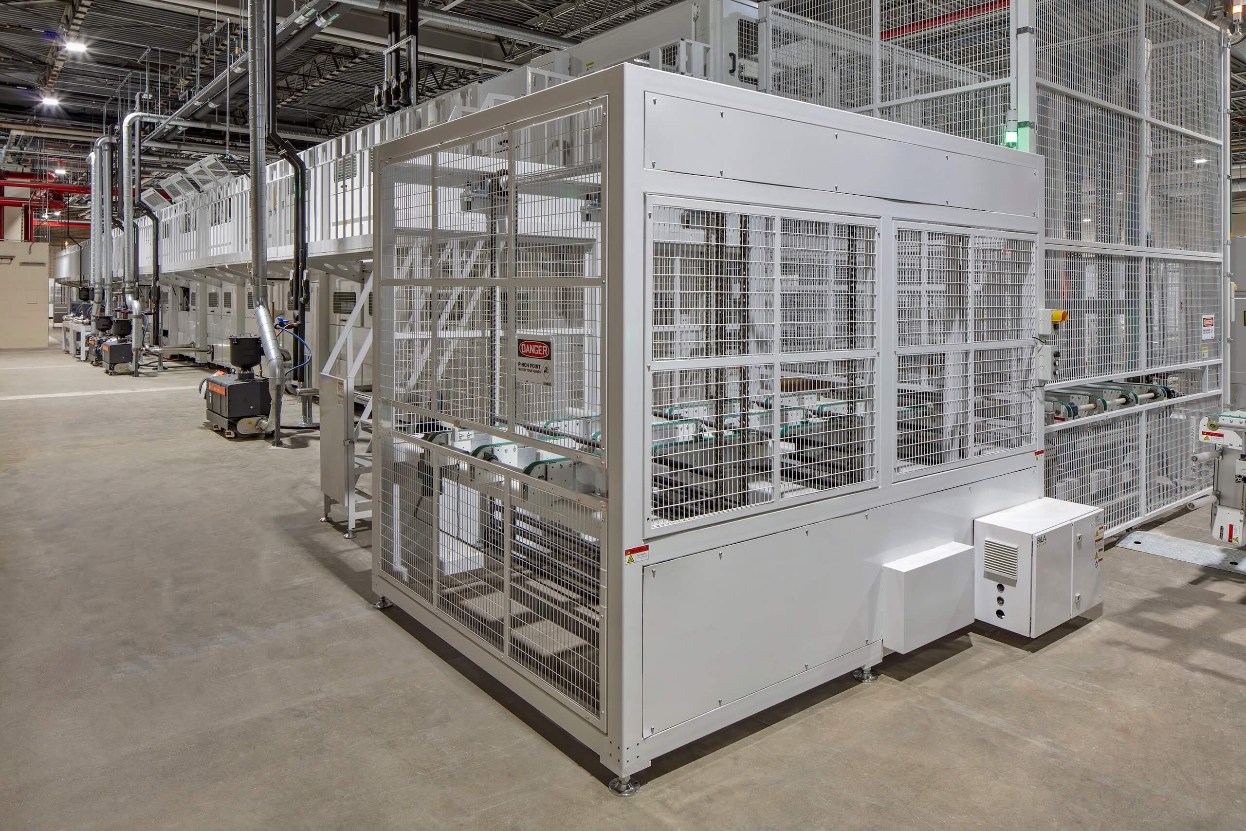 A large white industrial machine with metal grates stands in a spacious, clean factory. Conveyor belts and machinery are visible inside the enclosure, with more equipment and lighting in the background.