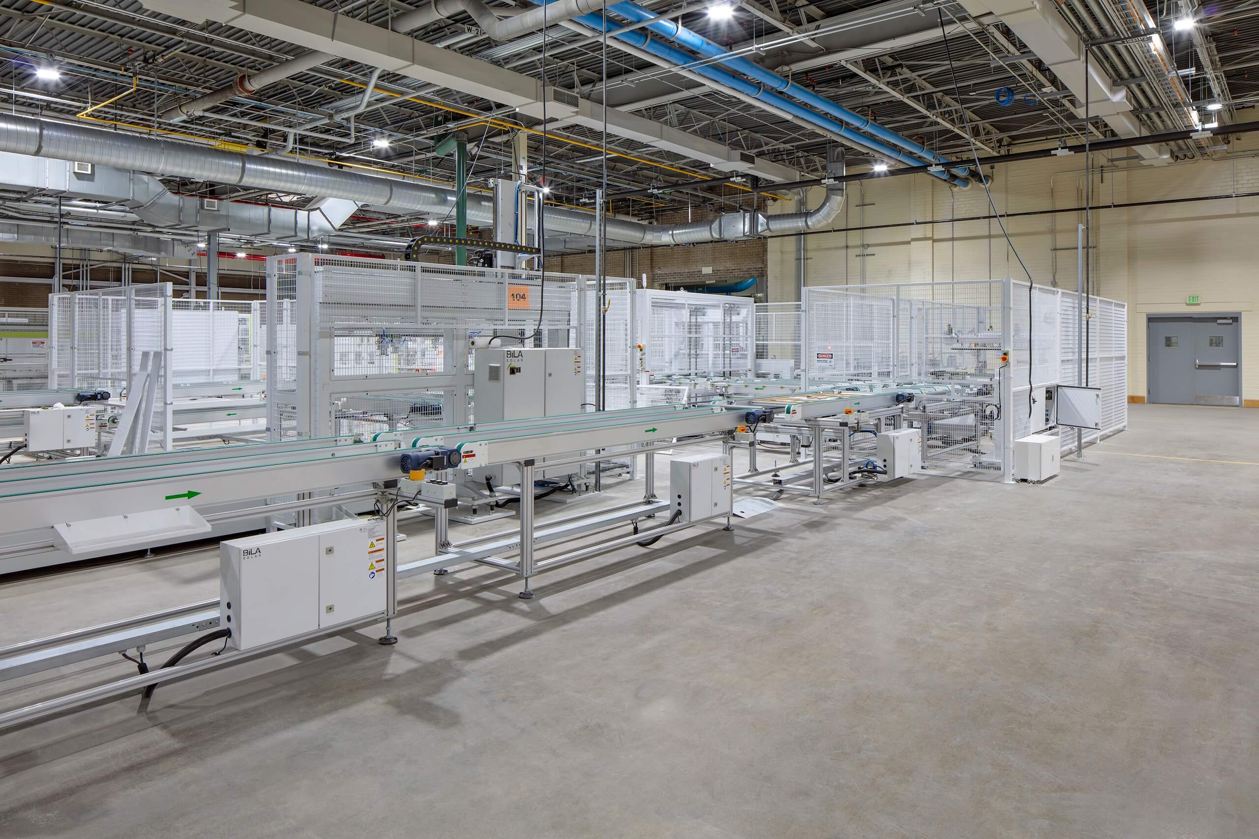 A brightly lit factory interior with metal conveyor belts, white machinery, and overhead pipes. The large room has a clean concrete floor and white mesh fencing around equipment.