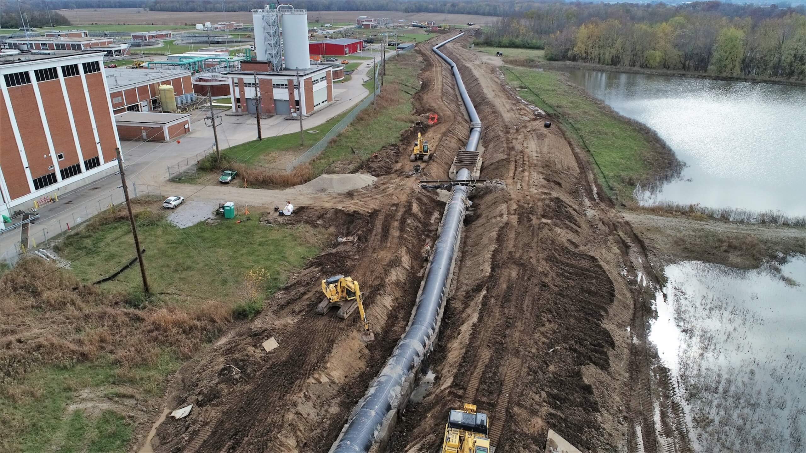 Aerial view of construction equipment installing a large pipeline next to a water treatment facility, with muddy ground and a pond nearby.