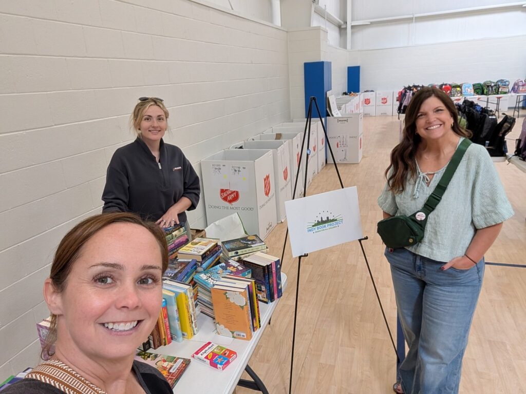 Three women stand by a table full of books in a gymnasium with donation bins in the background. Two women smile at the camera while one sorts books.
