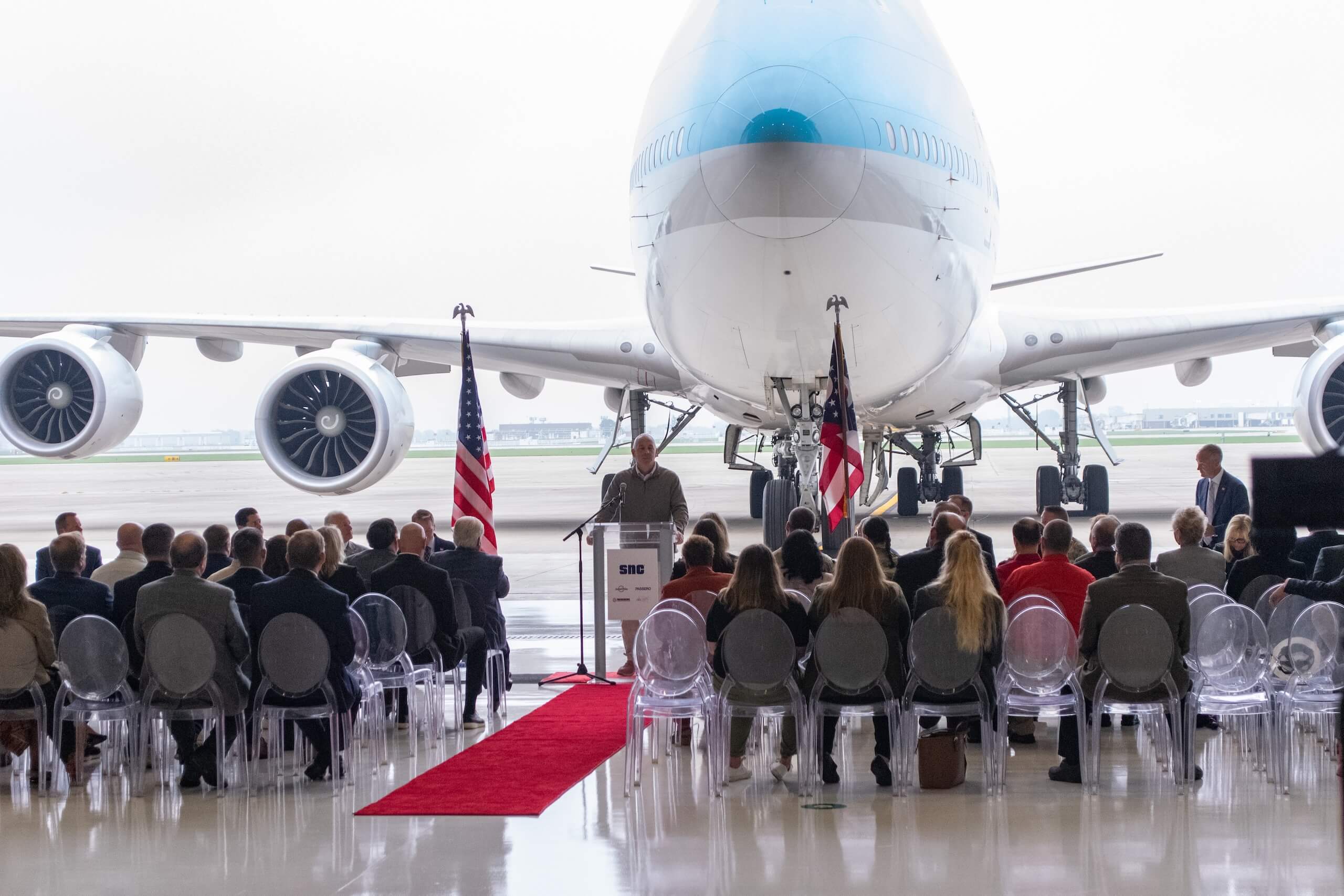 A speaker addresses a seated audience in front of a large airplane in a hangar. Two American flags flank the podium, and a red carpet leads to the stage.