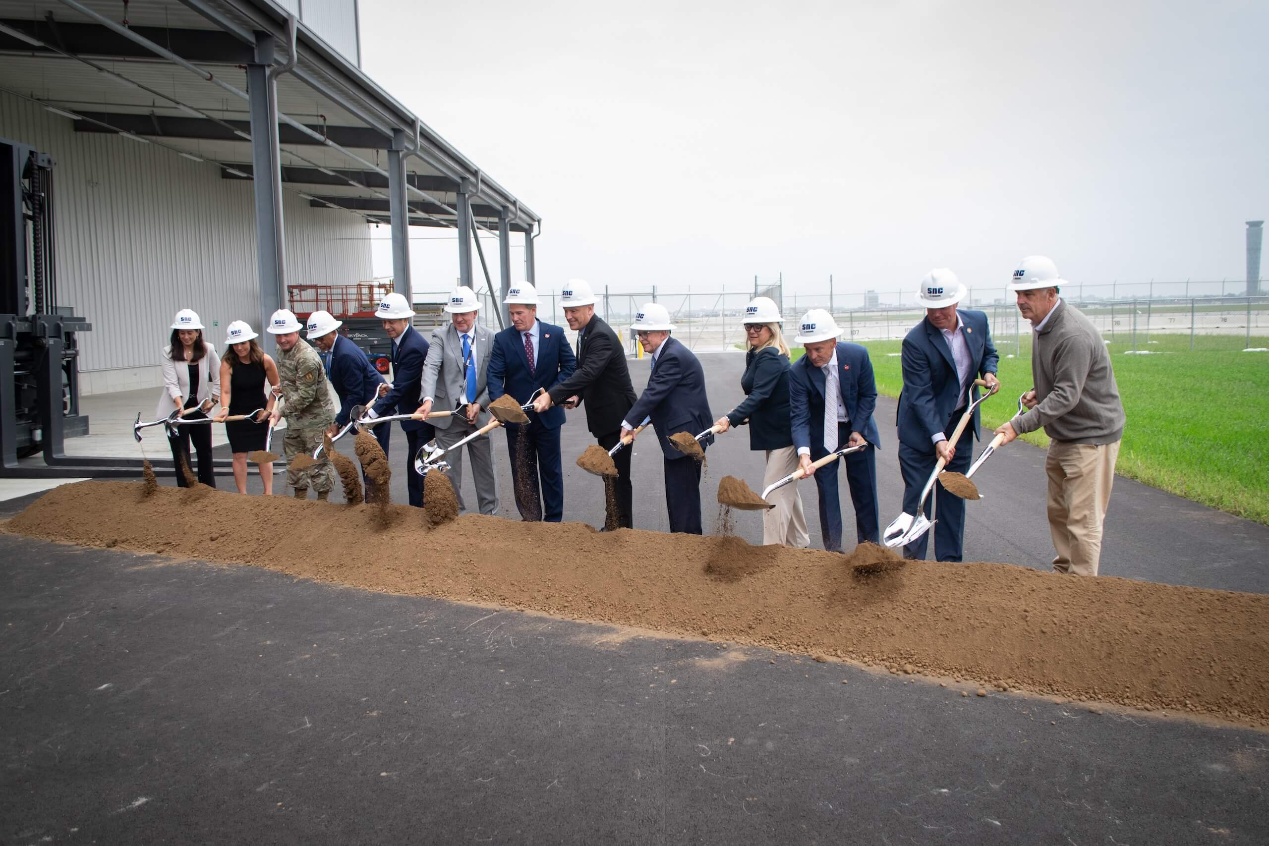 A group of people in hard hats and formal attire stand in a row, holding shovels and participating in a groundbreaking ceremony, turning over dirt on a construction site.