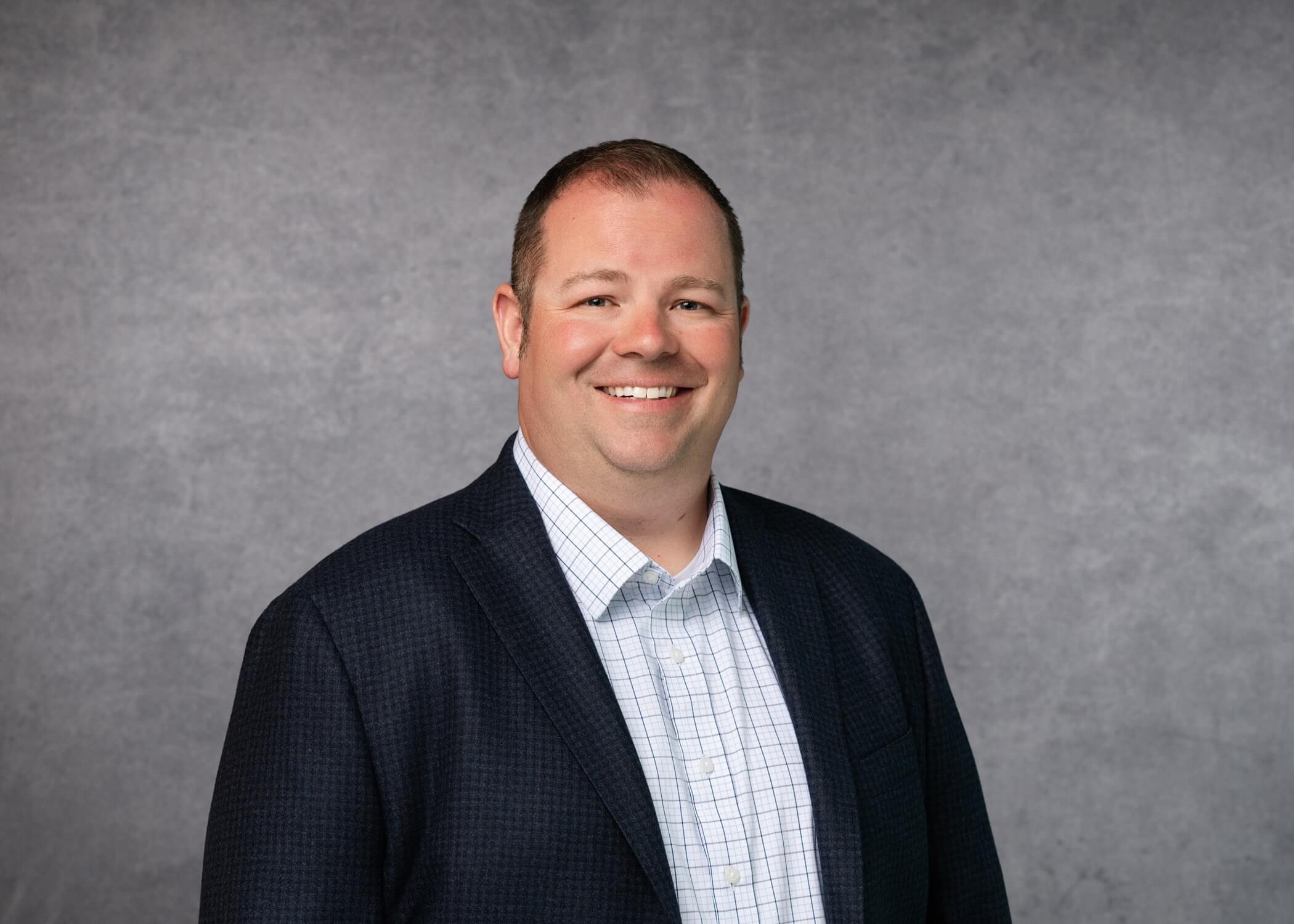 A man in a dark blazer and checked shirt smiles at the camera, standing in front of a neutral gray backdrop.