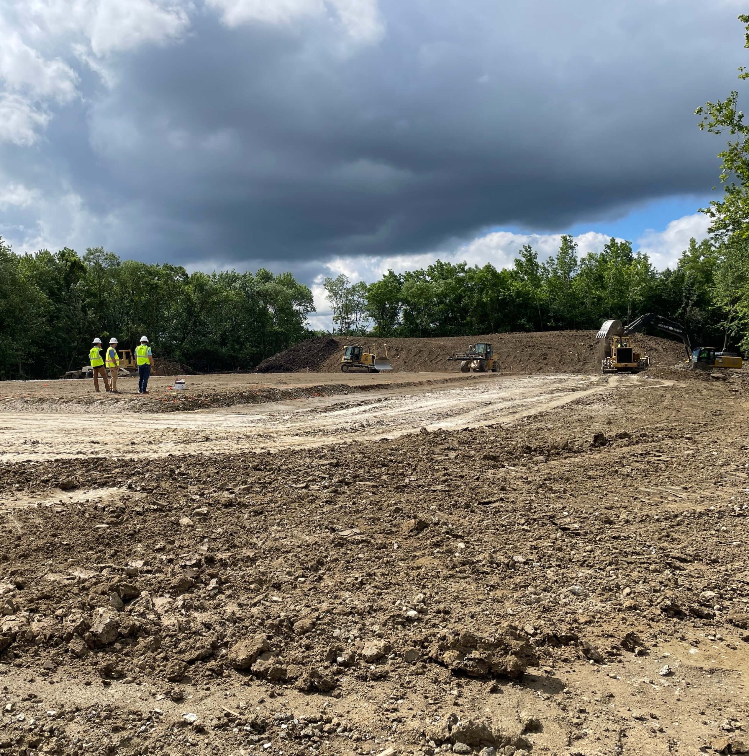 Several construction workers in safety vests stand on a dirt site with excavators. The sky is cloudy, and trees border the area.