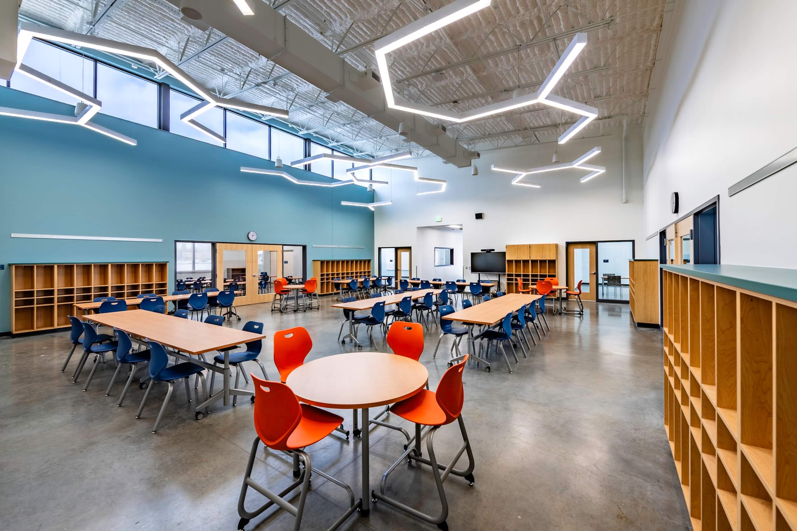 A modern school cafeteria with round and rectangular tables, orange and blue chairs, light blue walls, large windows, open shelves, and geometric ceiling lights. The room is clean, bright, and empty.