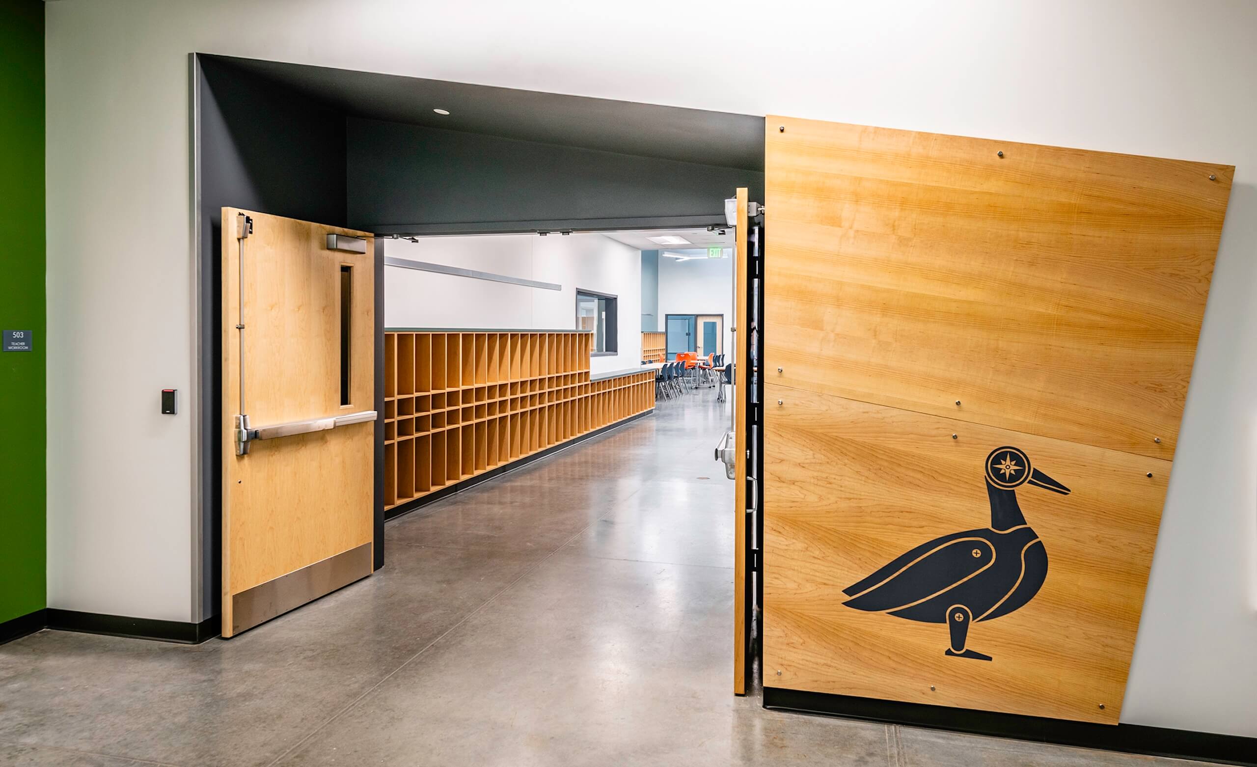 A hallway with wooden cubbies leads to a classroom with orange chairs. The open door has a wooden panel featuring a stylized bird illustration. The floor is polished concrete.